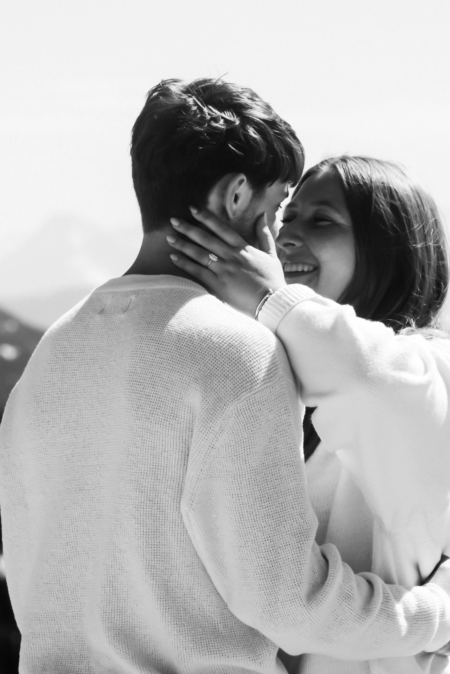A black and white photo of a couple embracing, with the woman holding the man's face and smiling, their noses touching.