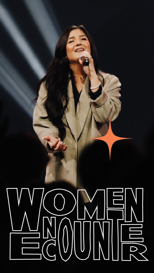 A woman singing into a microphone on stage during a Women in E-Count event, with a spotlight and dark background.