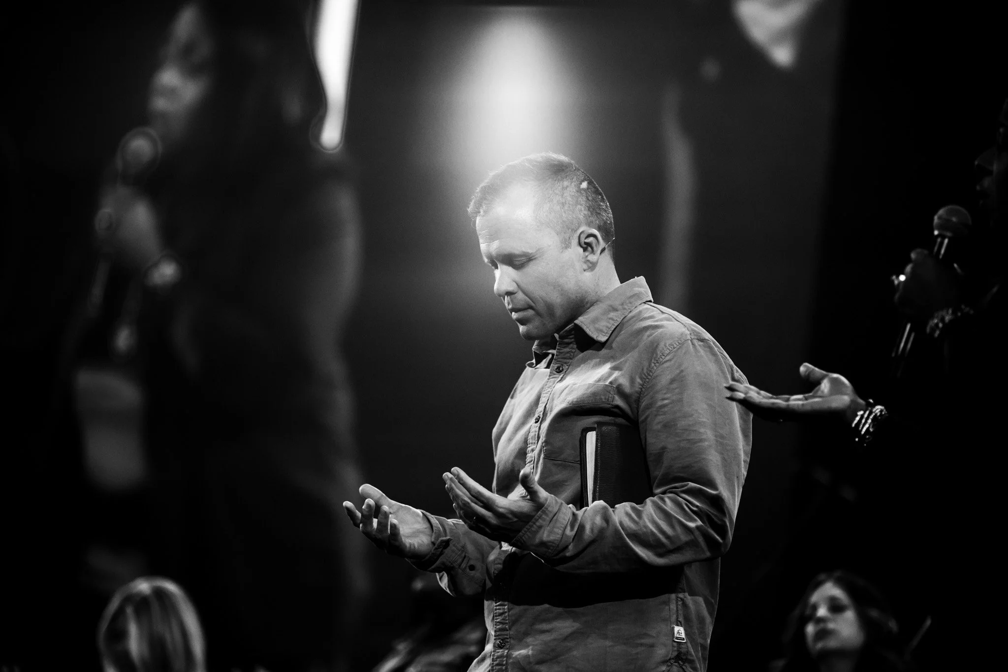 Black and white photo of a man with short hair and closed eyes, prayer hands, and a serious expression, surrounded by blurred people in a church or religious gathering, with soft backlighting.