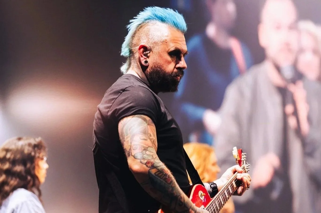 Man with blue mohawk hairstyle and a beard playing an electric guitar on stage, with a blurred group of people in the background.