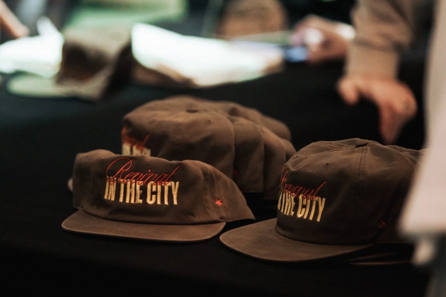 Several brown caps with embroidered text reading "Rejuvenated in the City" displayed on a black tablecloth, with a blurred background.
