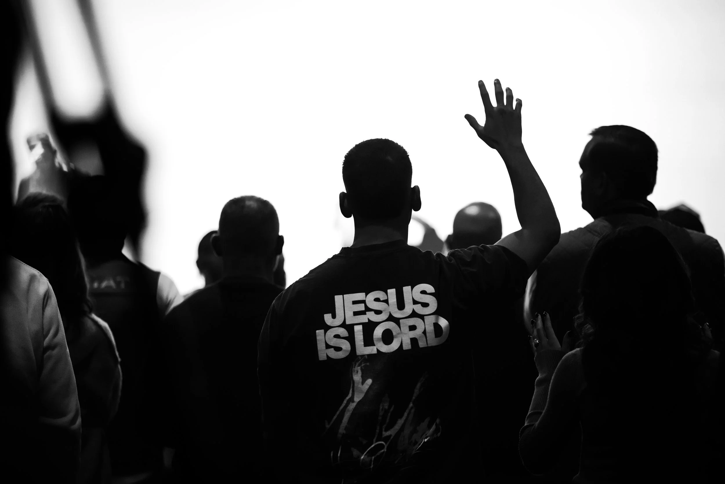 Black and white photo of a group of people with one person raising a hand. Someone is wearing a shirt with the text 'JESUS IS LORD' on the back.