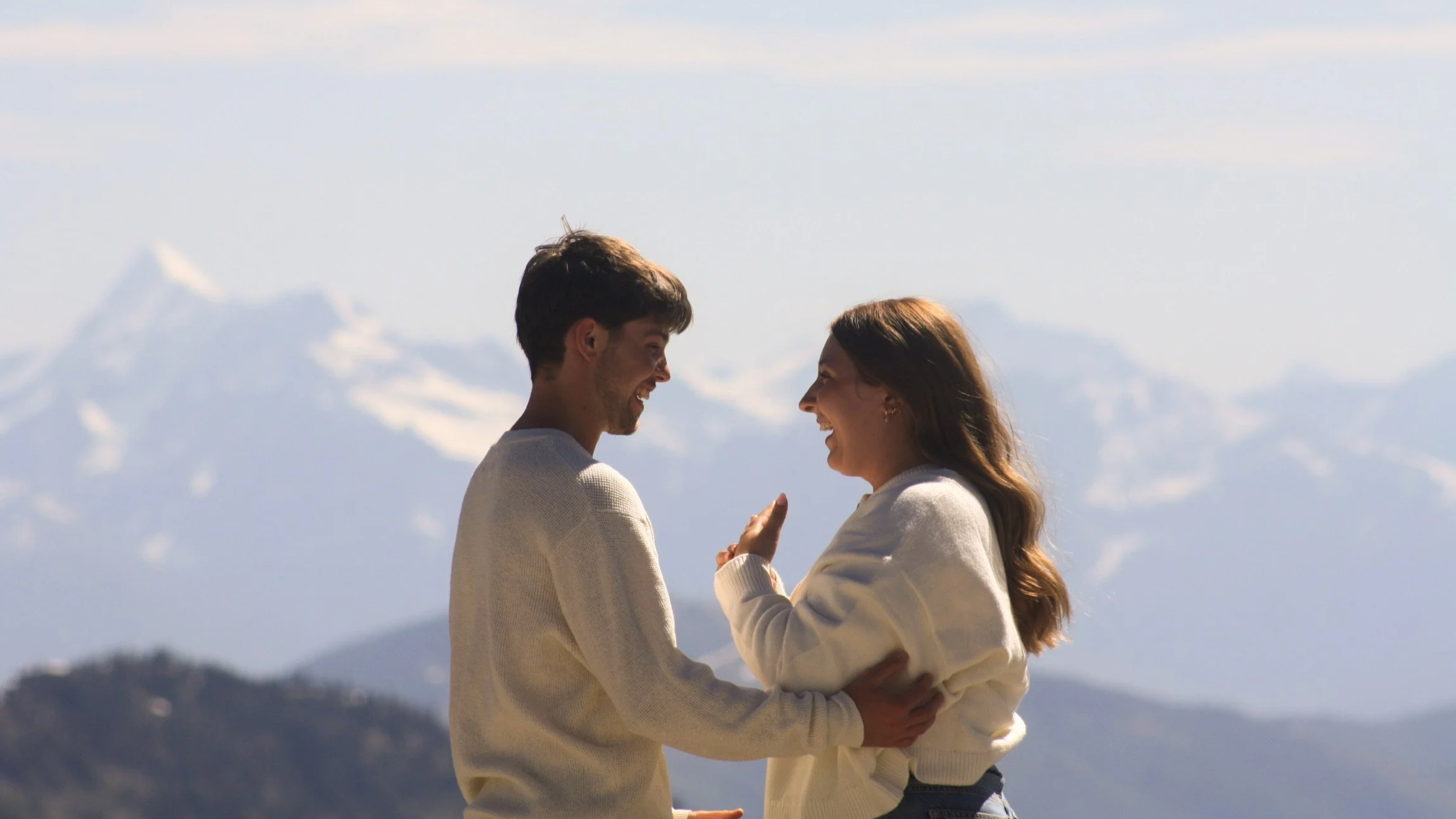 A young couple, a man and woman, smiling at each other while holding hands outdoors with snow-capped mountains in the background.