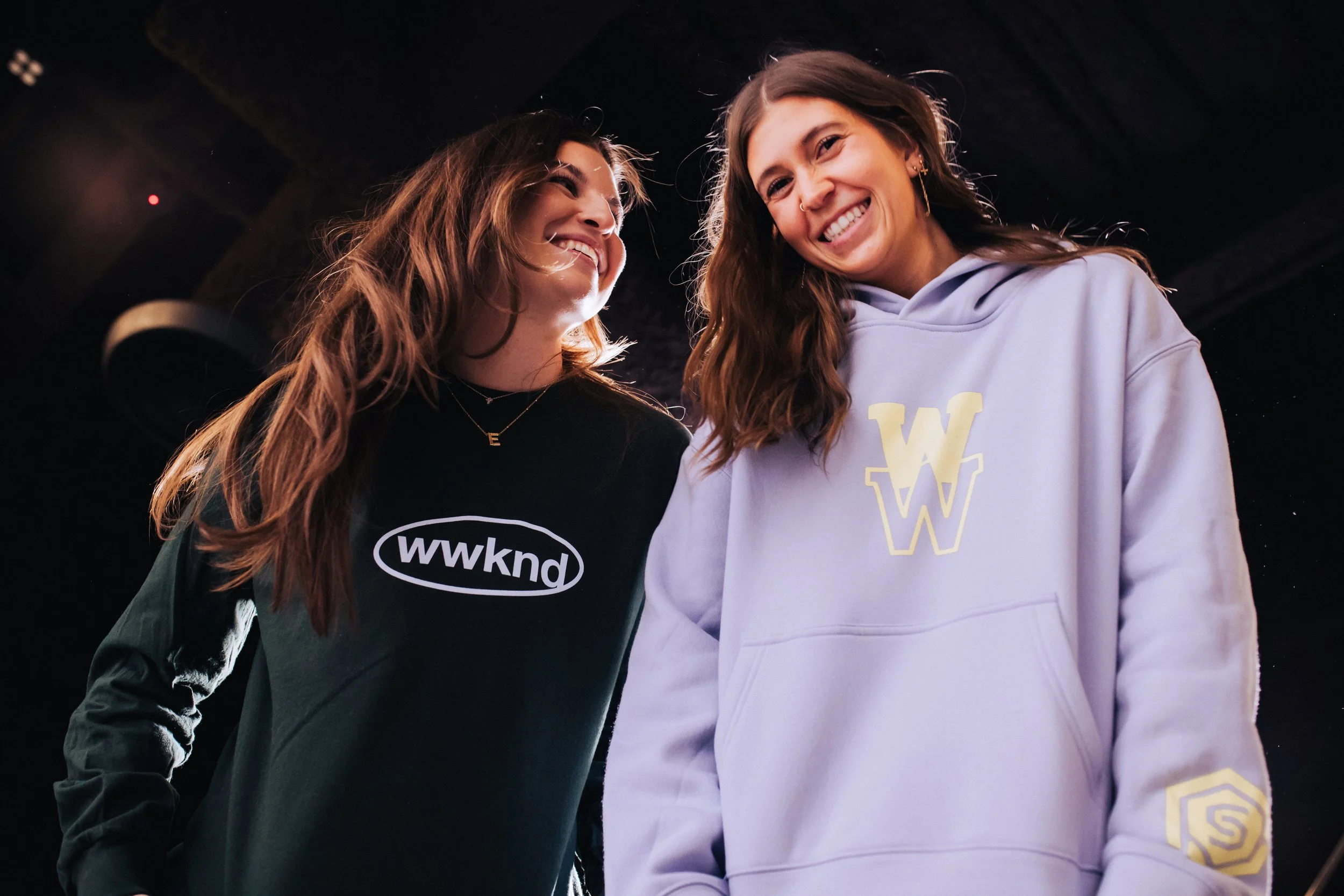 Two young women with long hair smiling and looking at each other, standing close together against a dark background, wearing casual sweatshirts.