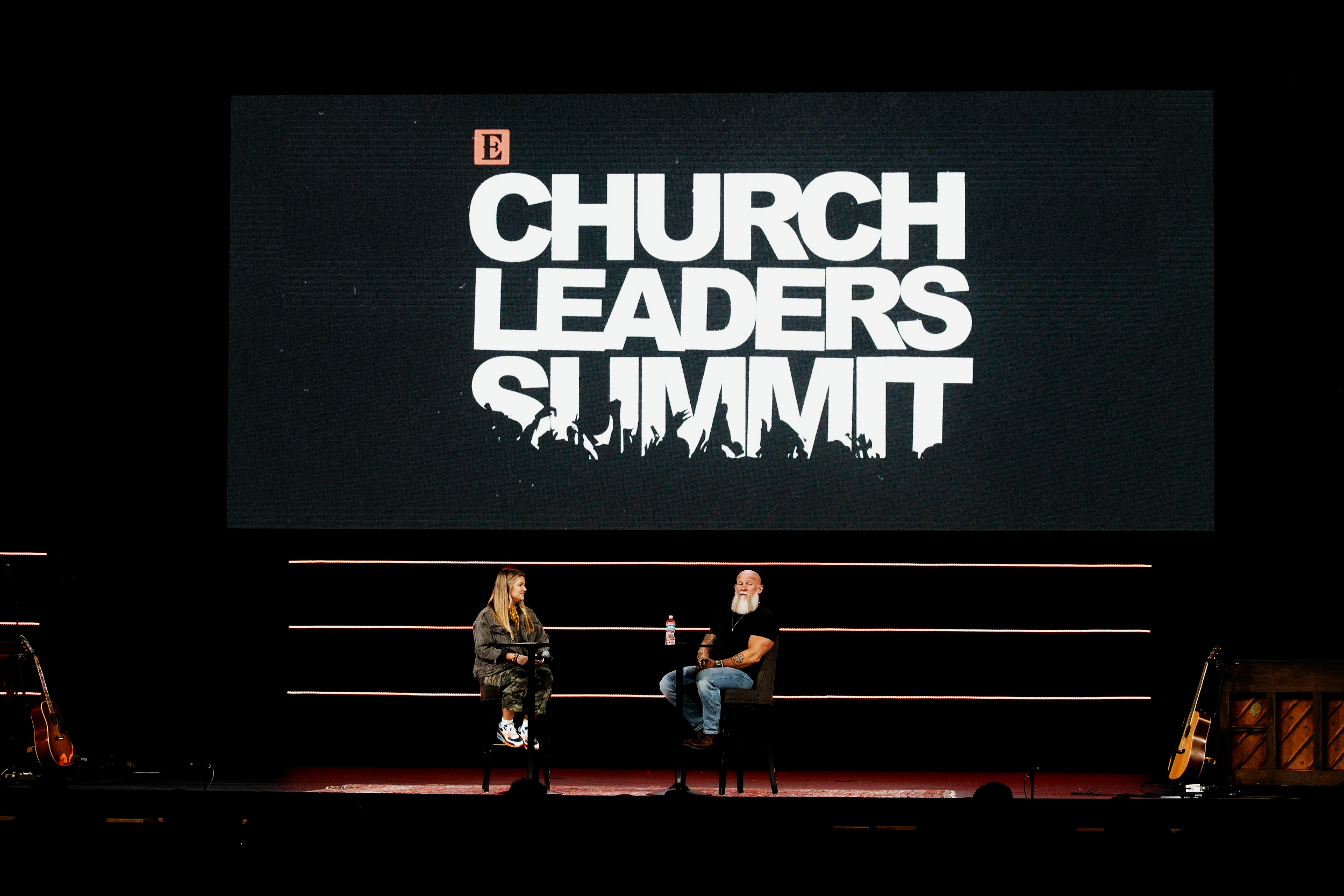 A large screen displays the words 'E Church Leaders Summit' in bold white text, with silhouettes of people raising their hands beneath. Two individuals are seated on stage below the screen, engaged in conversation, with guitars on stands and a water bottle on the table.