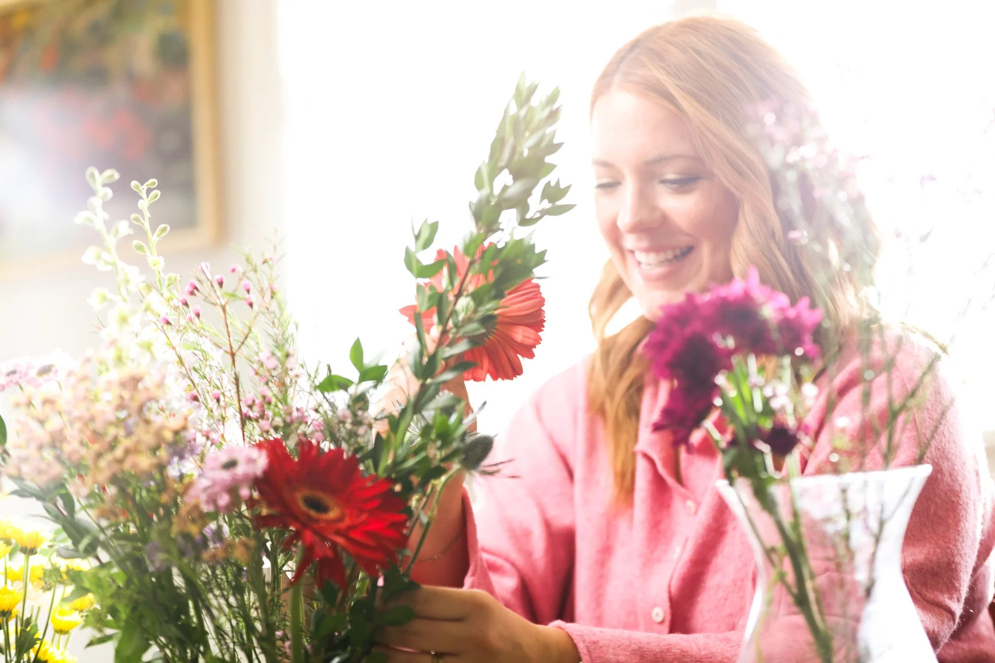 A woman with reddish hair wearing a pink sweater is smiling while looking at colorful flowers she is holding.