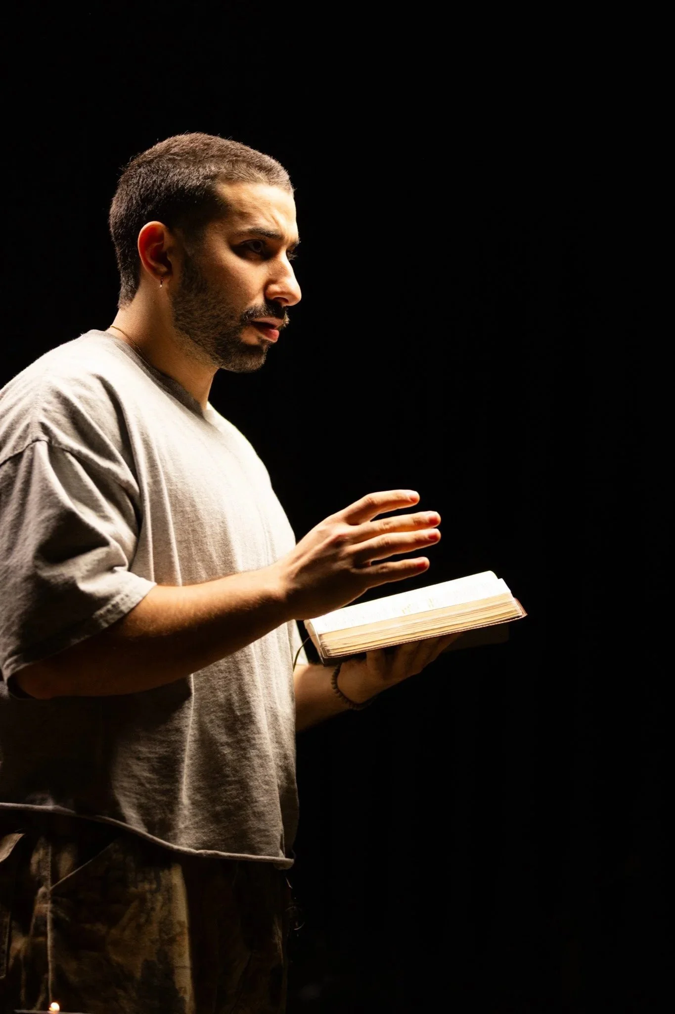 A man with short hair, a beard, and earrings is reading a book in a dark environment, illuminated only by a light source.