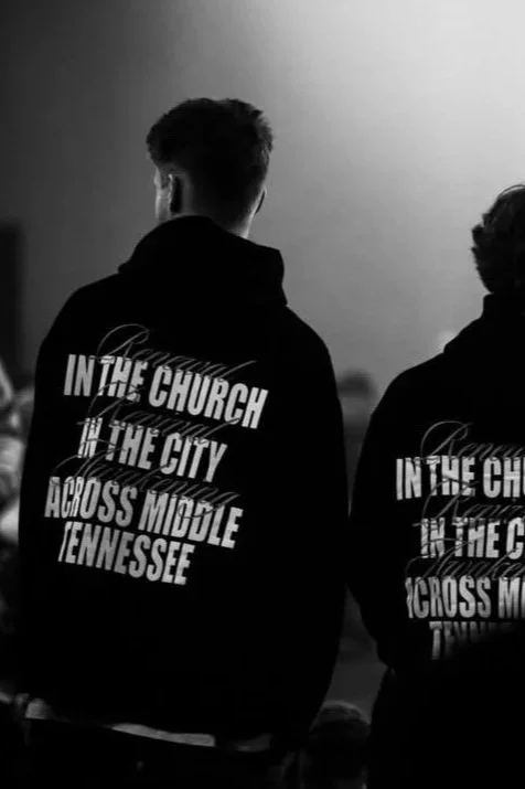 Two people wearing black hoodies with white text, facing away. The left hoodie says "IN THE CHURCH THE CITY ACROSS MIDDLE TENNESSEE."