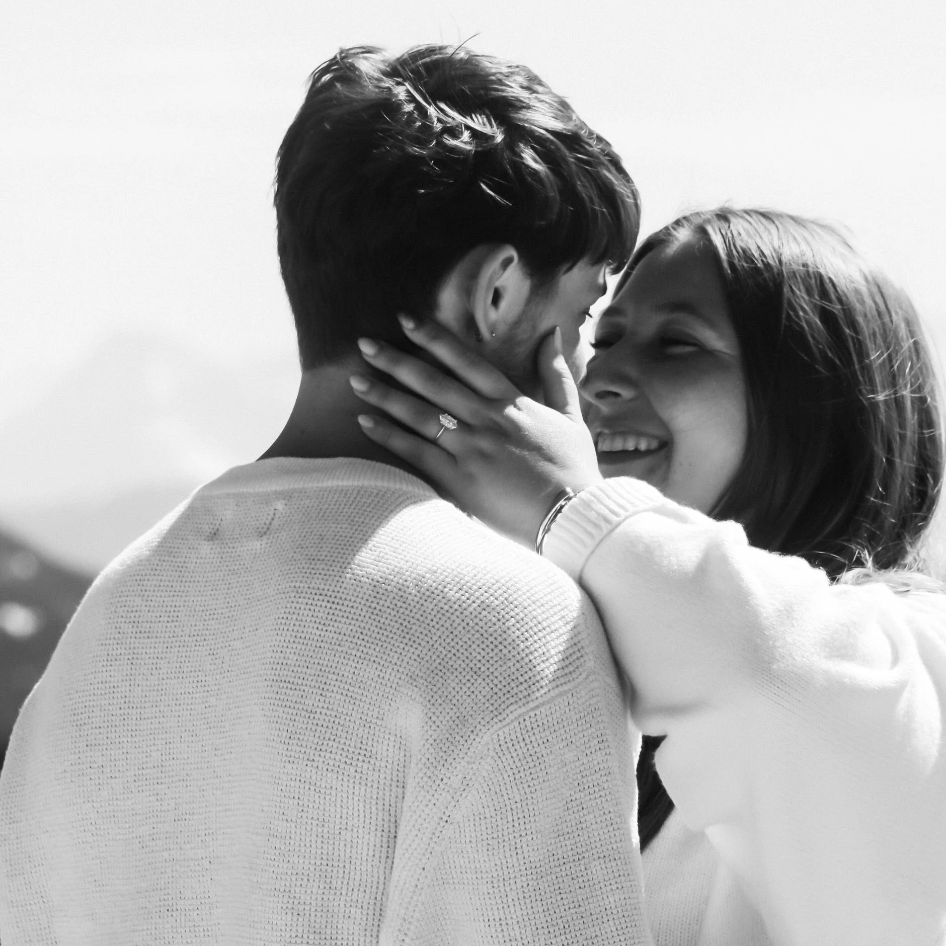 A black-and-white photograph of a couple sharing a kiss, with the woman touching the man's face and wearing a ring on her finger.