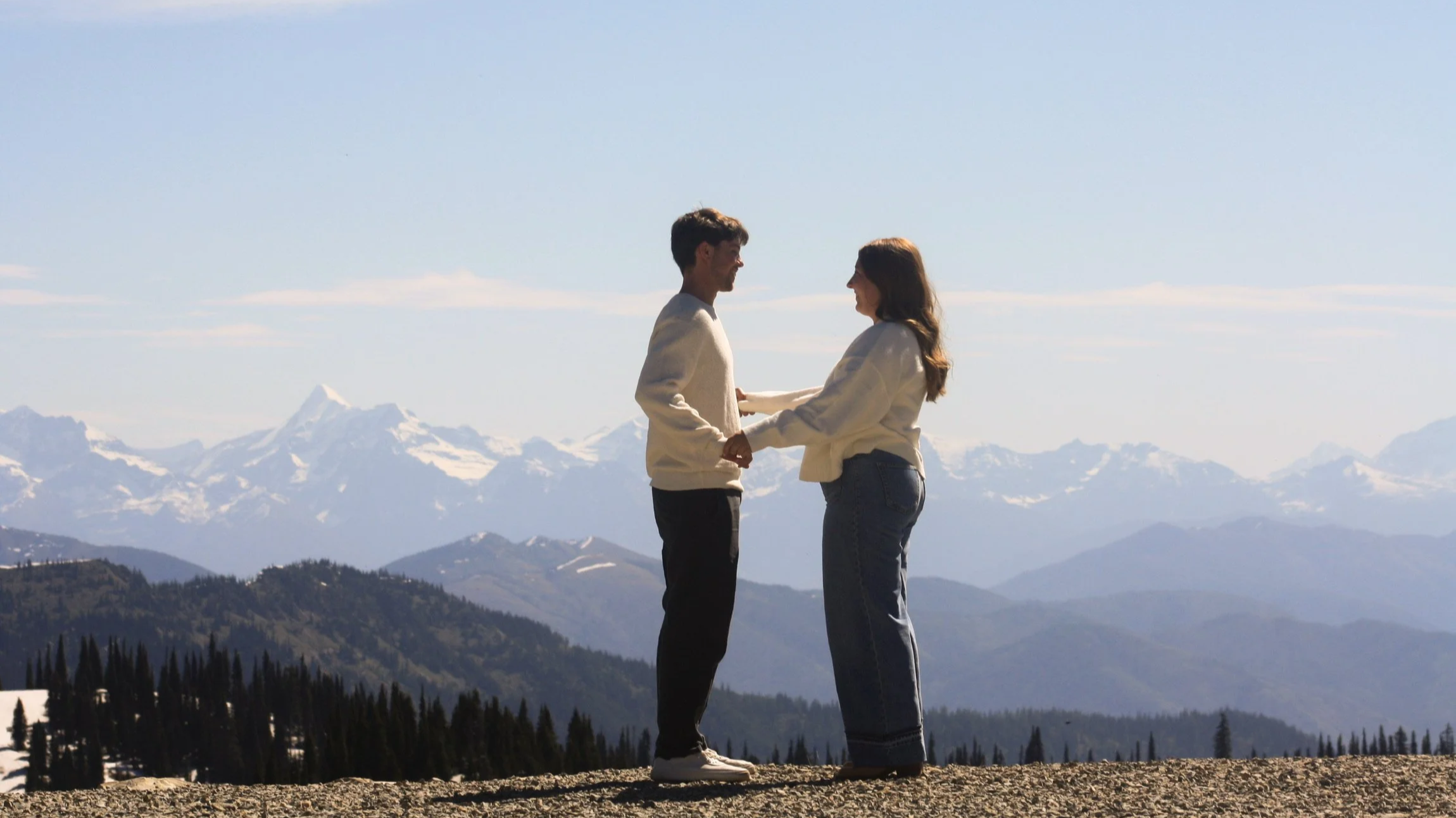 A couple holding hands and looking at each other in front of snow-capped mountains under a clear blue sky.