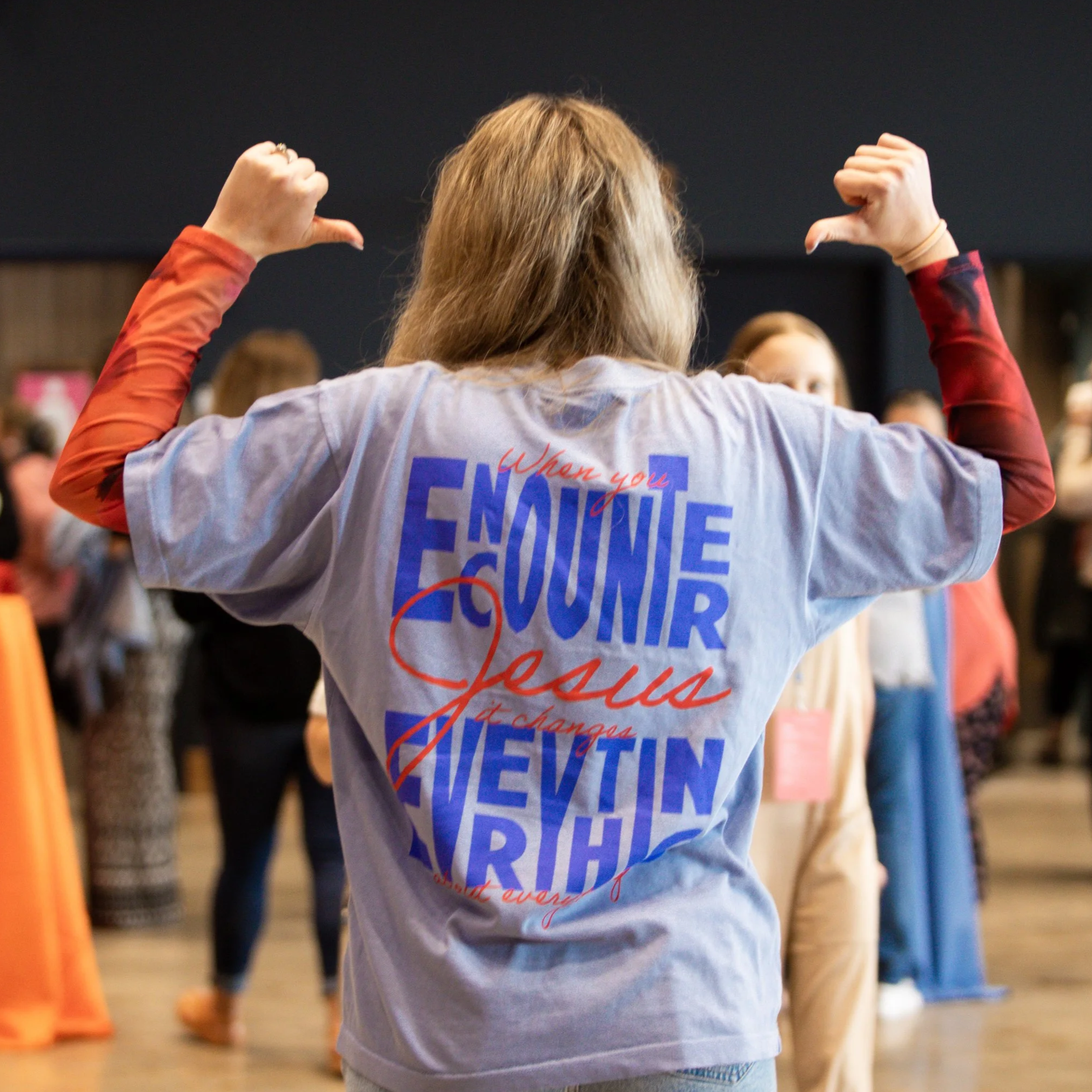 Back of a woman with long brown hair, wearing a gray shirt with writing, raising both fists in a crowd or gathering.