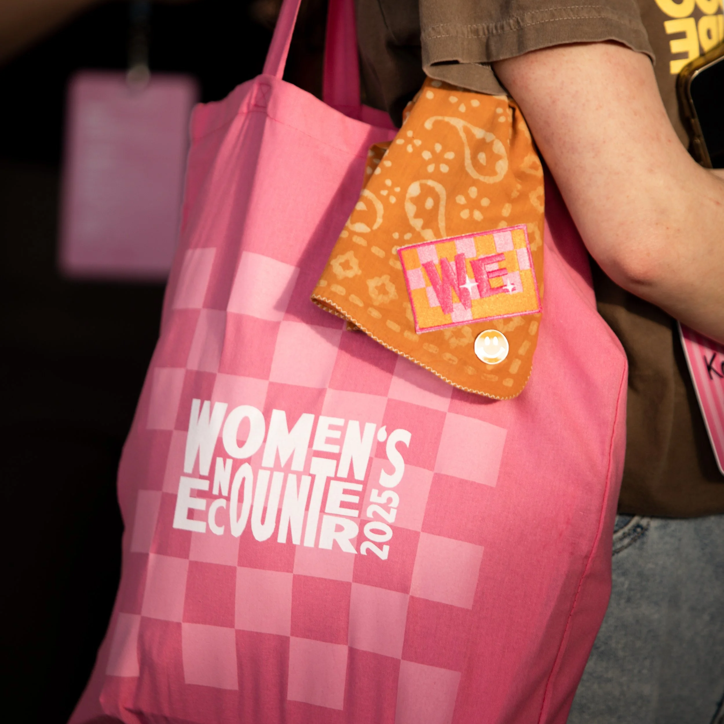 Close-up of a pink tote bag and an orange bandana with white patterns, worn over a person's shoulder and arm, partially covering a pink and white sign with white text that reads “WOMEN’S ELECTIONS”.