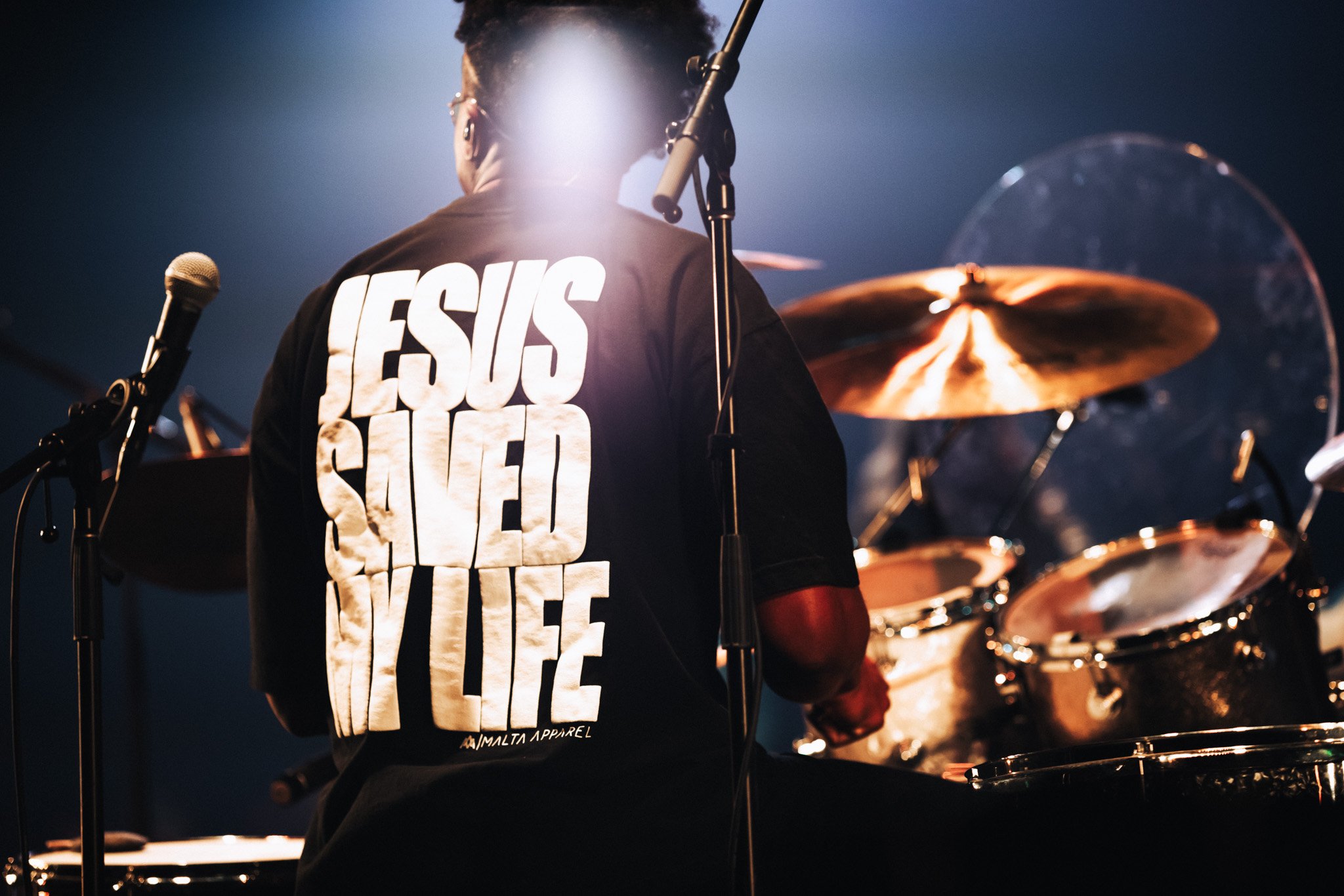 A musician playing drums on stage, wearing a black t-shirt with white text that reads 'JESUS SAVES' in large letters, with additional smaller text underneath, during a concert or performance.