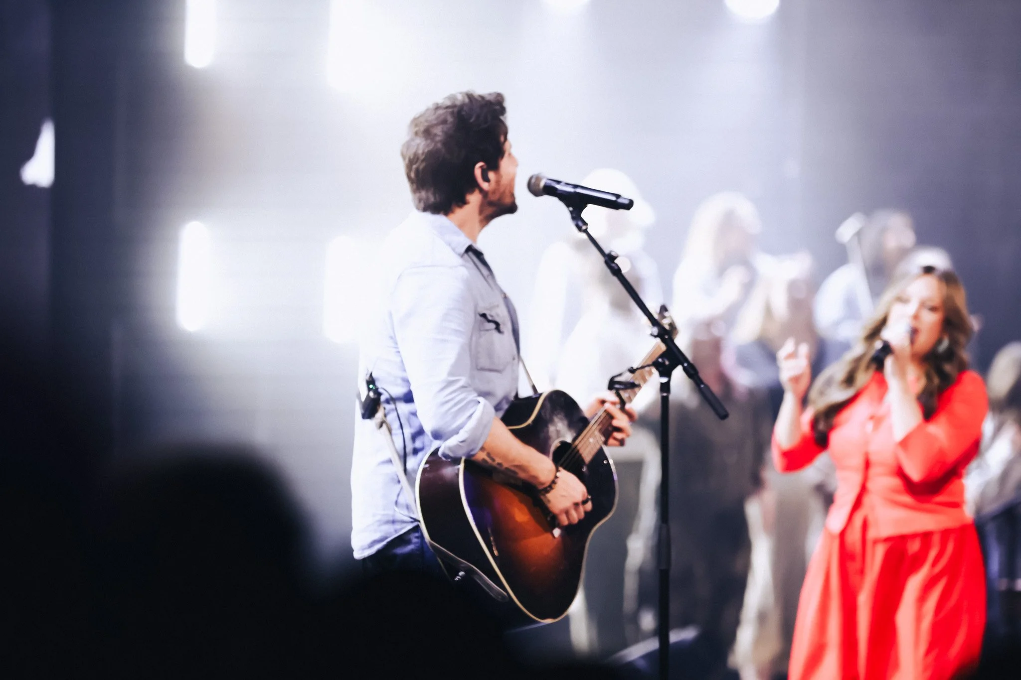 A man playing an acoustic guitar and singing into a microphone on stage, with women singing in the background, in a brightly lit indoor setting.
