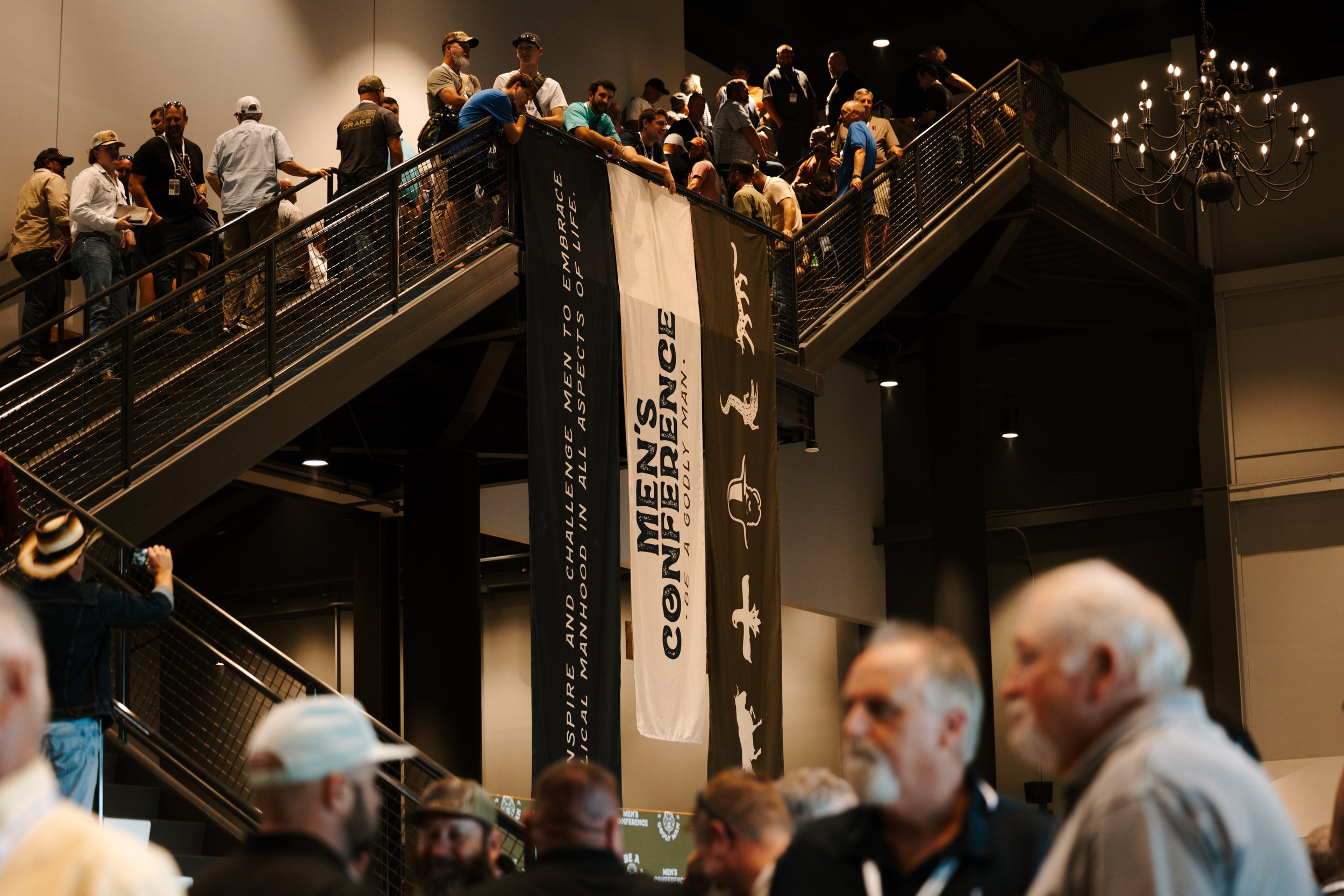 People line up on stairs at a conference, with banners for the Modern Men’s Conference and a chandelier hanging from the ceiling.