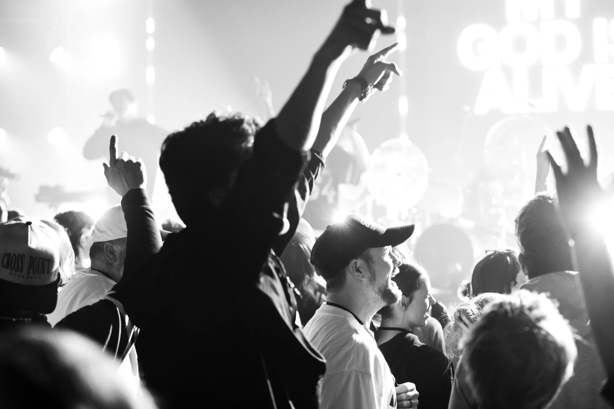 Black and white photo of a crowd at a concert or event, with some people raising their hands and wearing hats, and a bright light in the background.