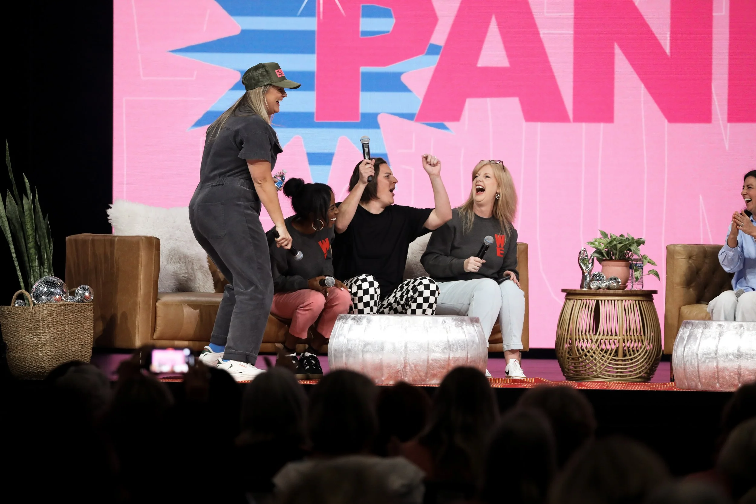 Group of women sitting on a stage, laughing and chatting, with some raising their hands in a friendly manner, during a panel or event with a pink and blue background.