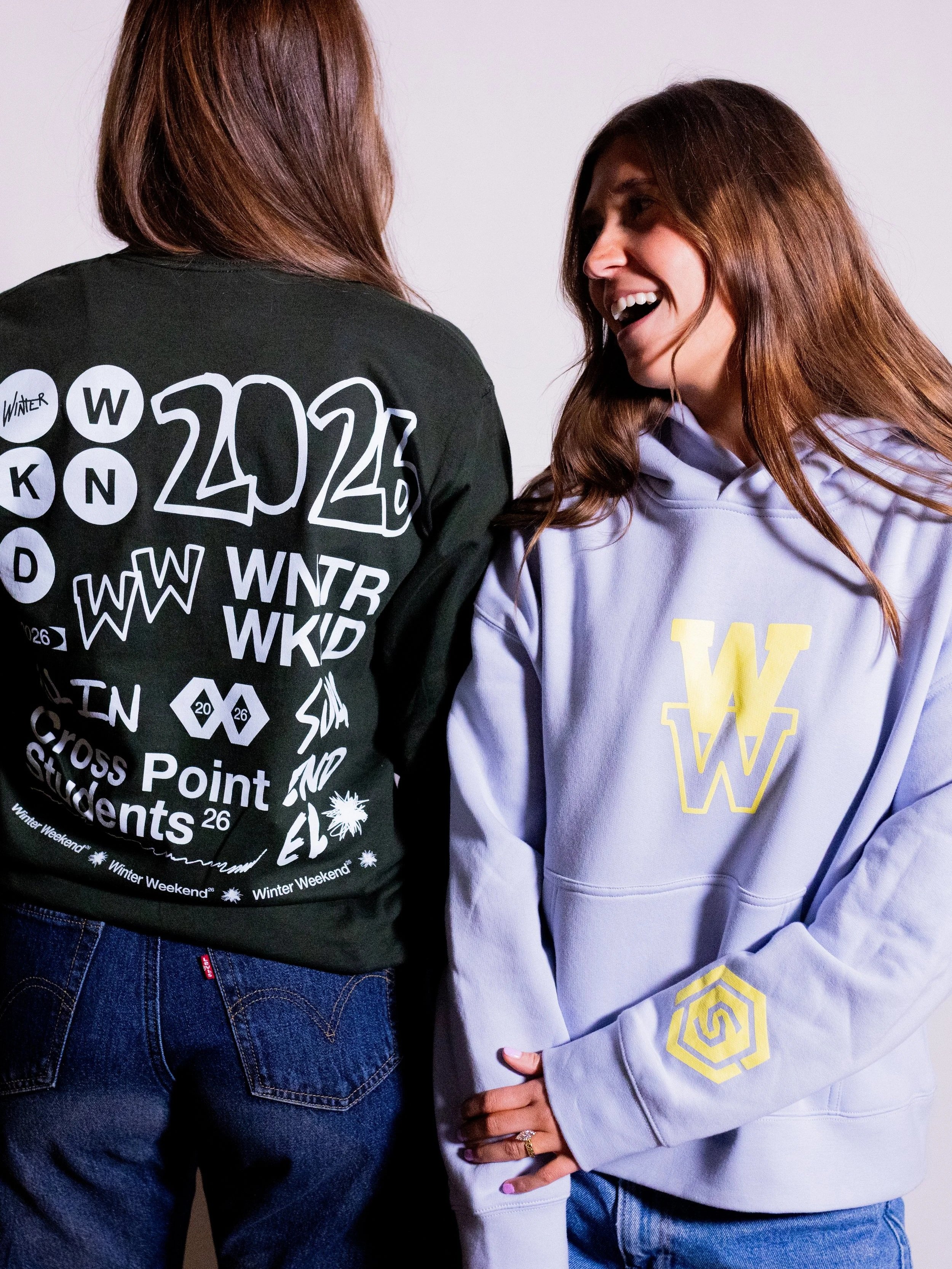 Two women standing close together, one with back facing forward wearing a black sweatshirt with white graphics, the other facing sideways wearing a light purple hoodie with yellow emblem, smiling and engaging in conversation.