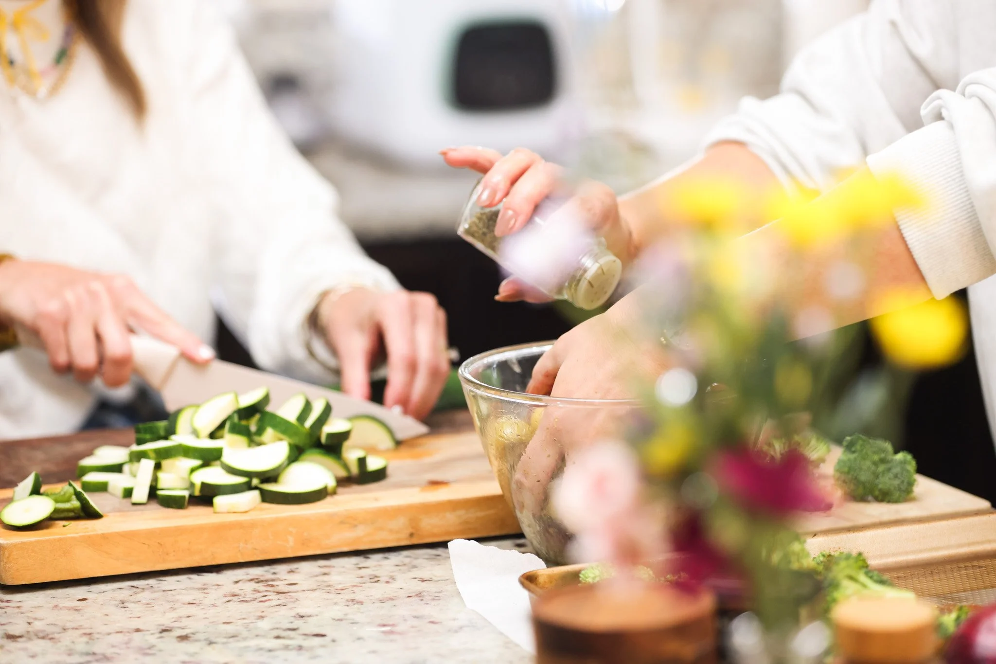 People preparing vegetables like zucchini on a kitchen counter with a glass bowl, surrounded by bowls of herbs and flowers.