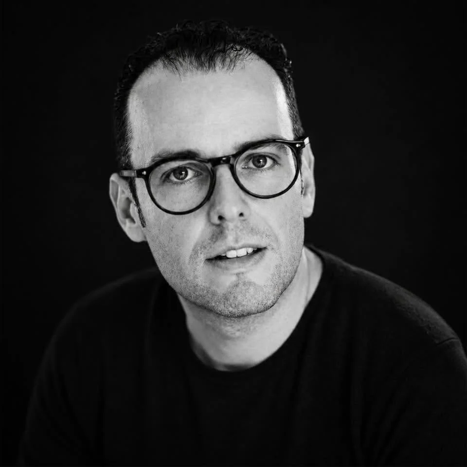 Retrato en blanco y negro de un hombre con gafas y cabello oscuro, usando una camiseta negra, con fondo negro.