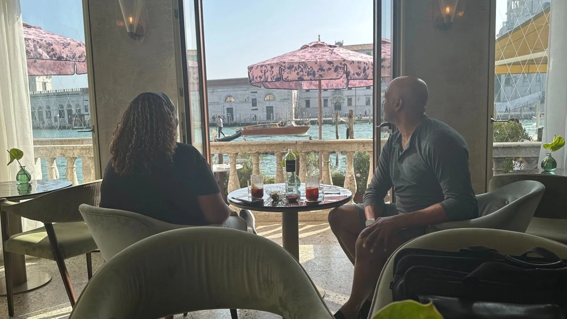 Two people sitting at a round table inside a cafe near a window, overlooking a canal with boats and historic buildings, umbrellas outside, and potted plants inside.