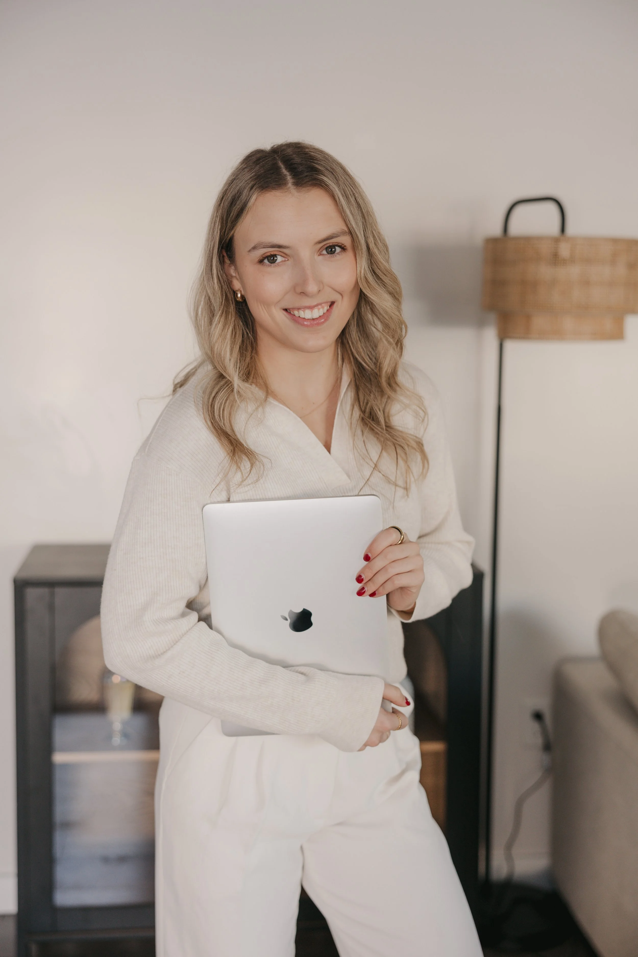 A woman with wavy blonde hair, wearing a cream-colored sweater and white pants, smiling and holding a silver laptop with an Apple logo, in a modern indoor setting with a wicker lampshade and minimal decor.