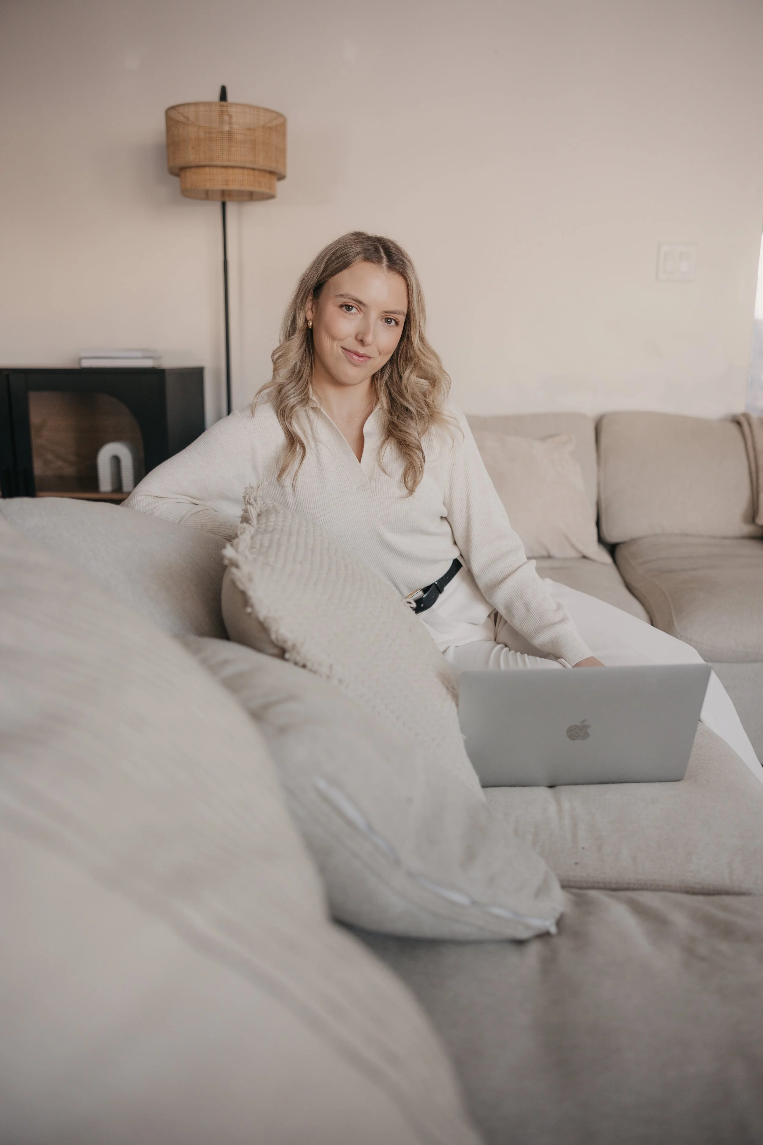 A woman with wavy blonde hair sitting on a beige couch in a cozy living room, using a silver MacBook laptop on her lap. She is smiling and wearing a cream-colored top and white pants, with a background featuring a tall wicker lamp and a black TV stand.