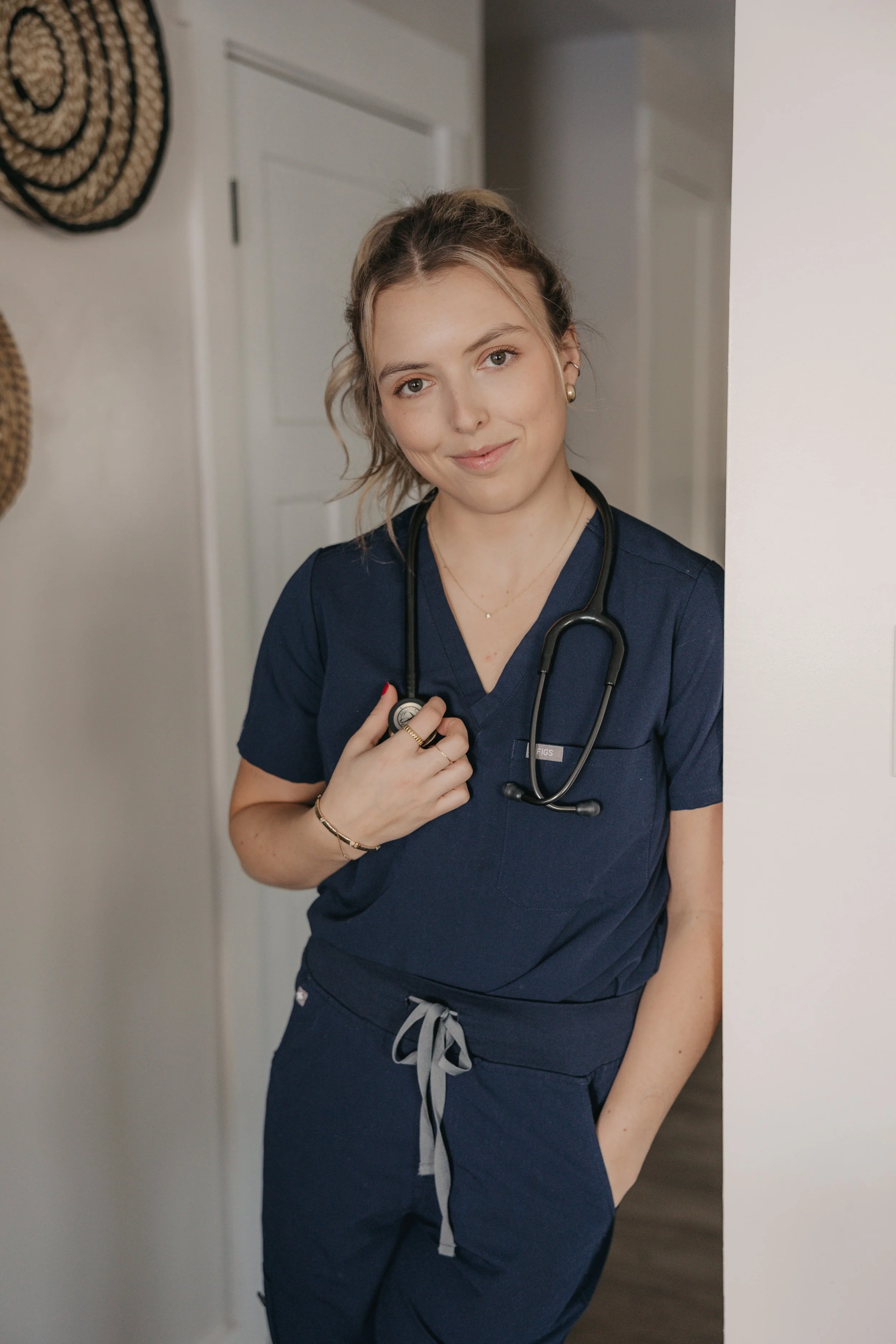 A young woman in navy scrubs with a stethoscope around her neck standing in a doorway.