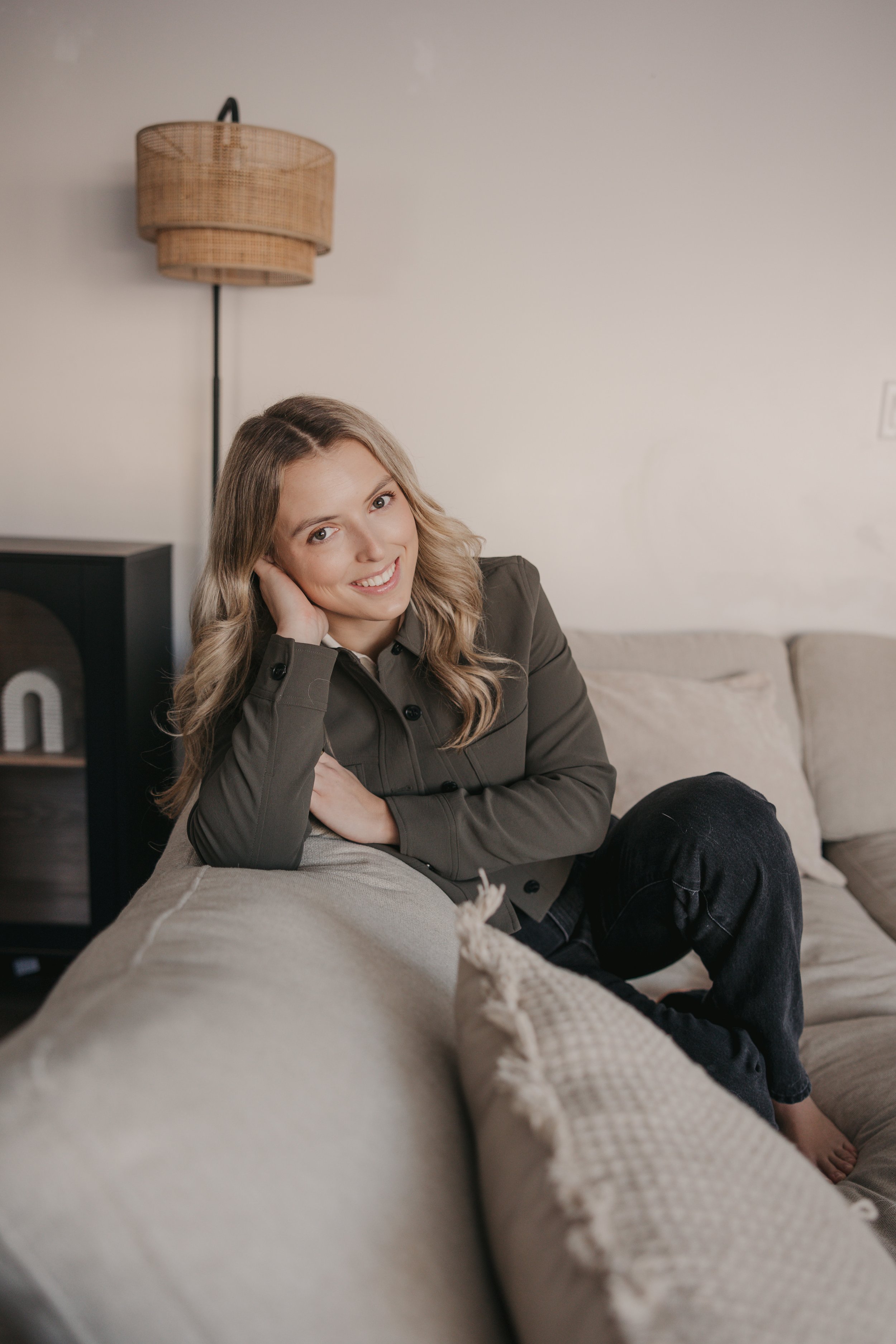 A woman with long wavy blonde hair sitting on a beige couch, smiling and resting her head on her hand. Behind her, there is a wall with a wicker lamp fixture and a black speaker or cabinet. She is wearing a dark green jacket and black pants.