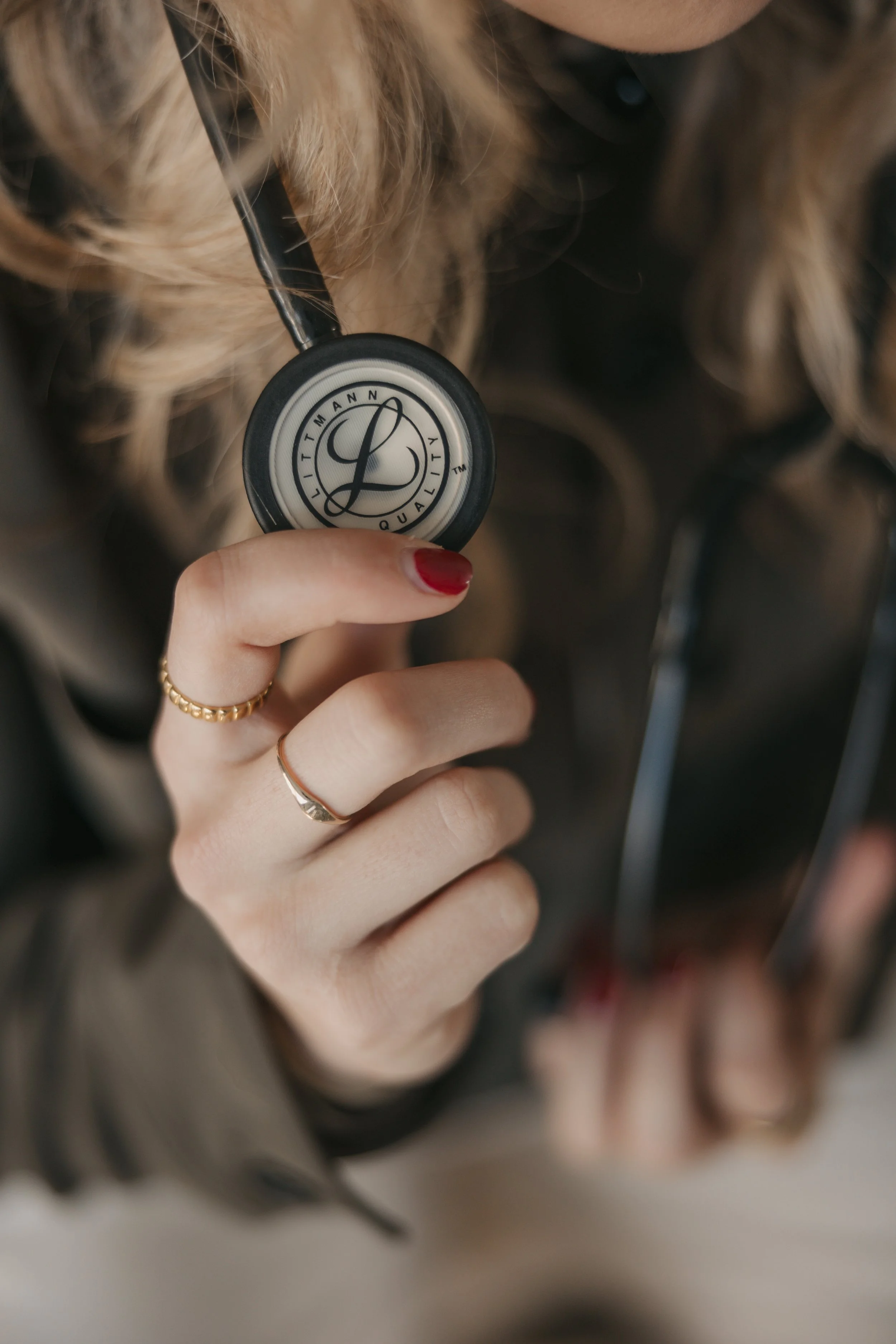 Close-up of a hand holding a stethoscope with a logo on it, featuring a stylized letter L and the words Ultimate Quality, with long, blonde, curly hair in the background.