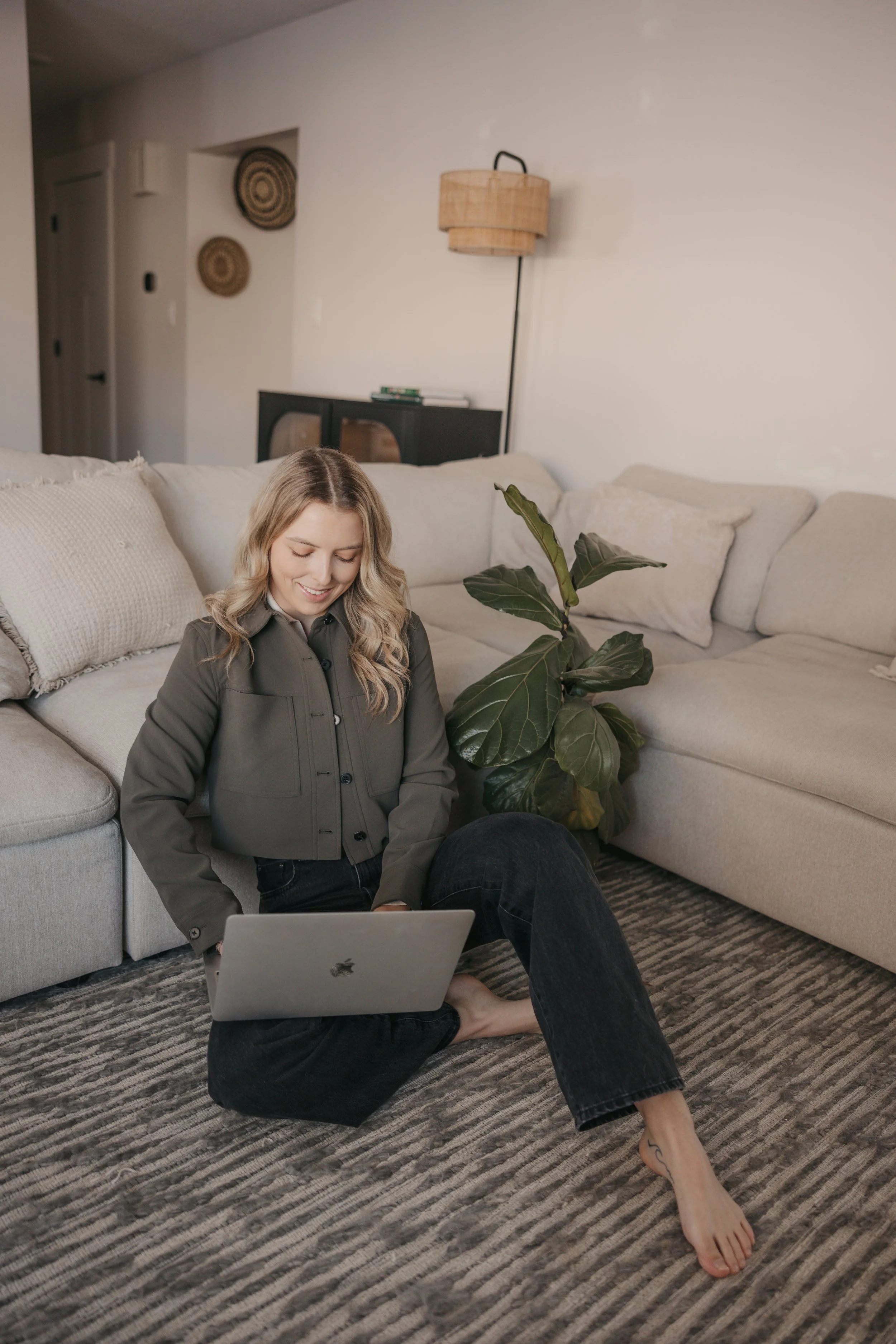 A young woman with blonde hair and a tattoo on her foot is sitting on the floor in a living room, smiling at her laptop. She is wearing a dark jacket and jeans. There is a beige sofa, a large green plant, a black cabinet, and decorative wall art in the background.
