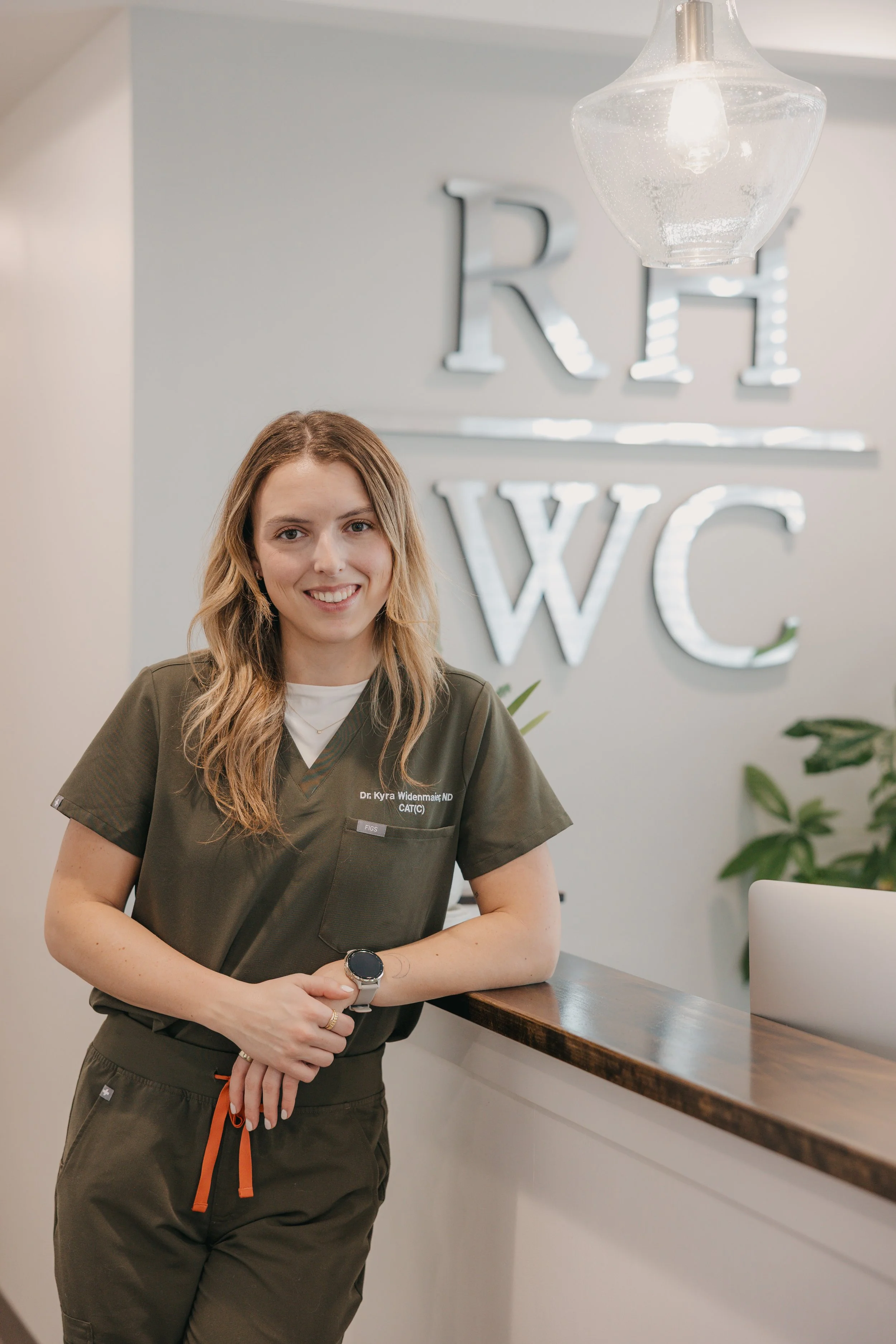 A young woman with long, wavy hair smiling stands in front of a wall with large silver letters spelling out 'ER' and 'WC'. She is wearing medical scrubs with a name tag that reads 'Dr. Kyra Widenmaier ND' and a smartwatch, with her arms resting on a wooden counter next to a laptop and some green plants.