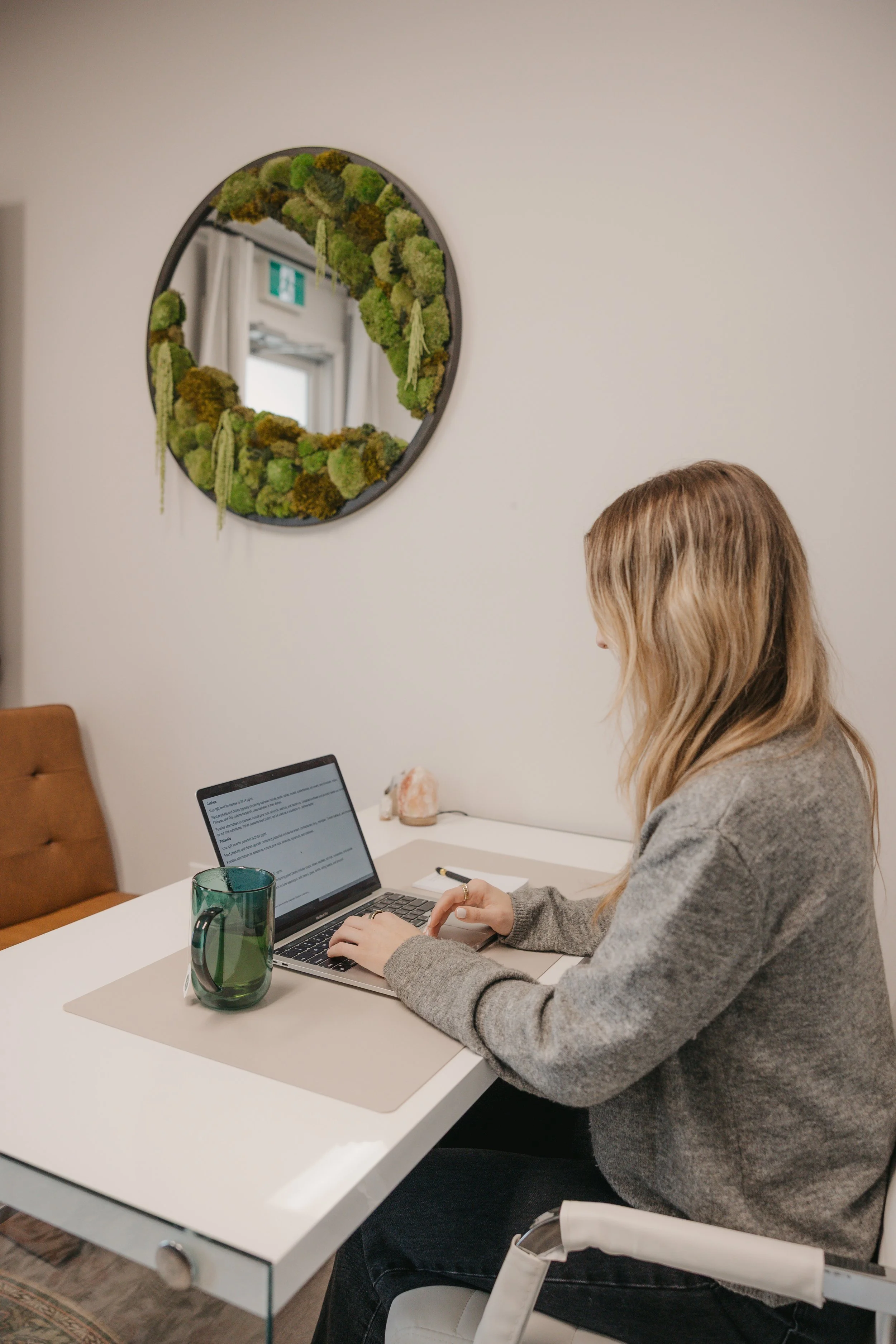 A woman sitting at a white desk working on a laptop in a room with a mirror decorated with green moss on the wall. A green glass mug, a notepad, and a pen are on the desk.