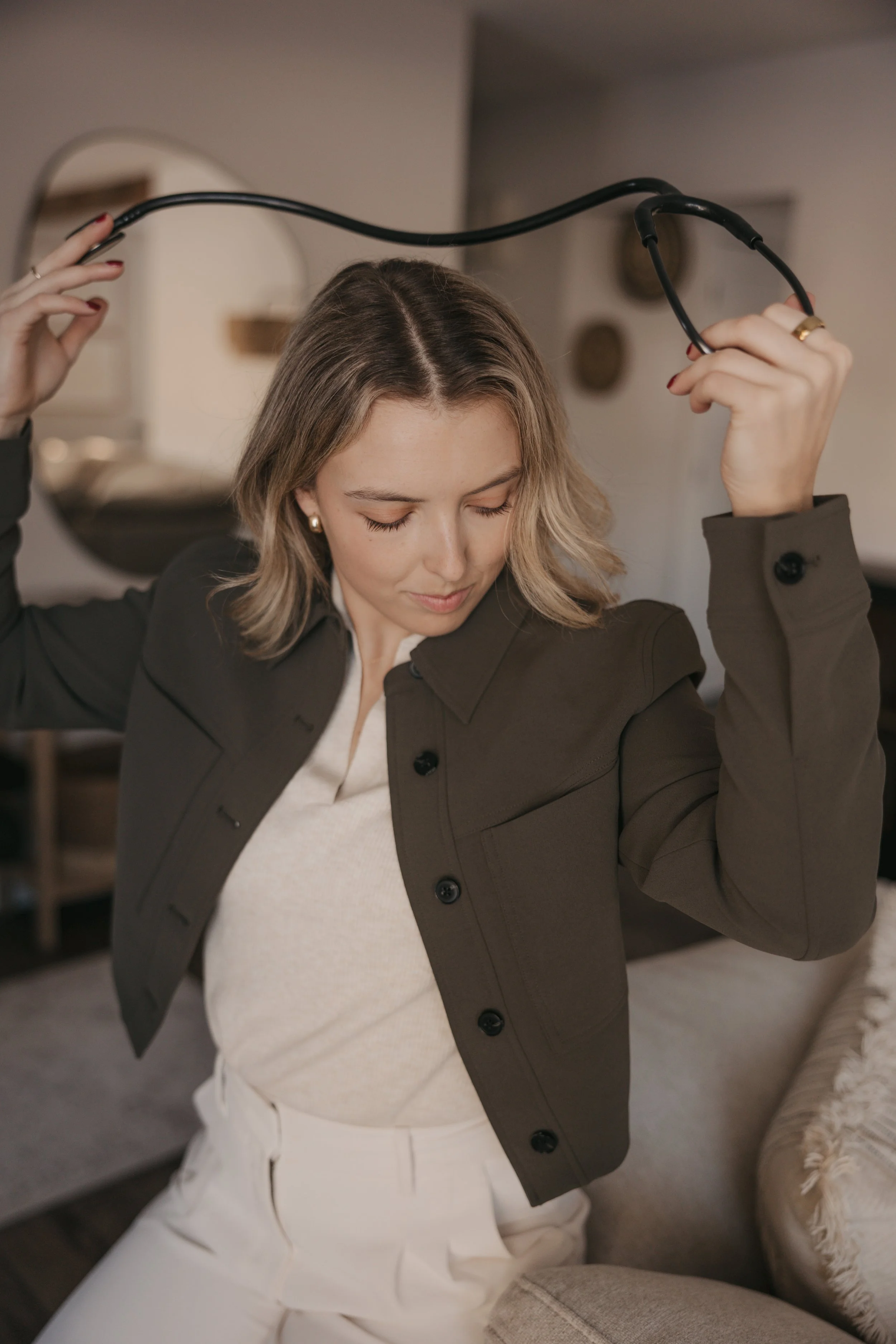 A woman with blonde hair, closed eyes, and a slight smile, holding a black cable above her head with both hands. She is wearing a dark blazer, a white sweater, and white pants, sitting in a cozy indoor setting.