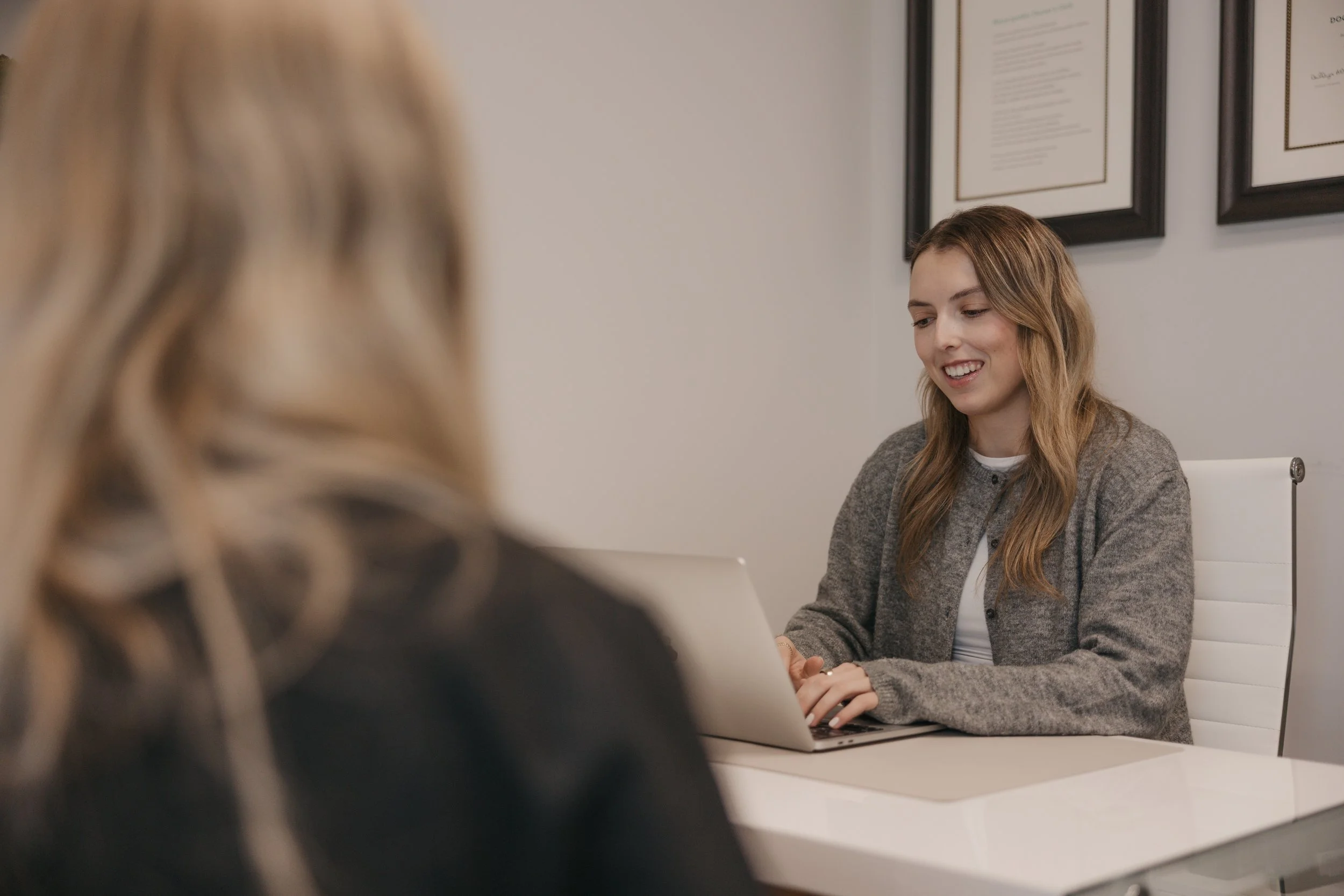 A woman with long brown hair and a gray sweater smiling while working on a laptop at a white table in an office, with framed documents on the wall behind her.