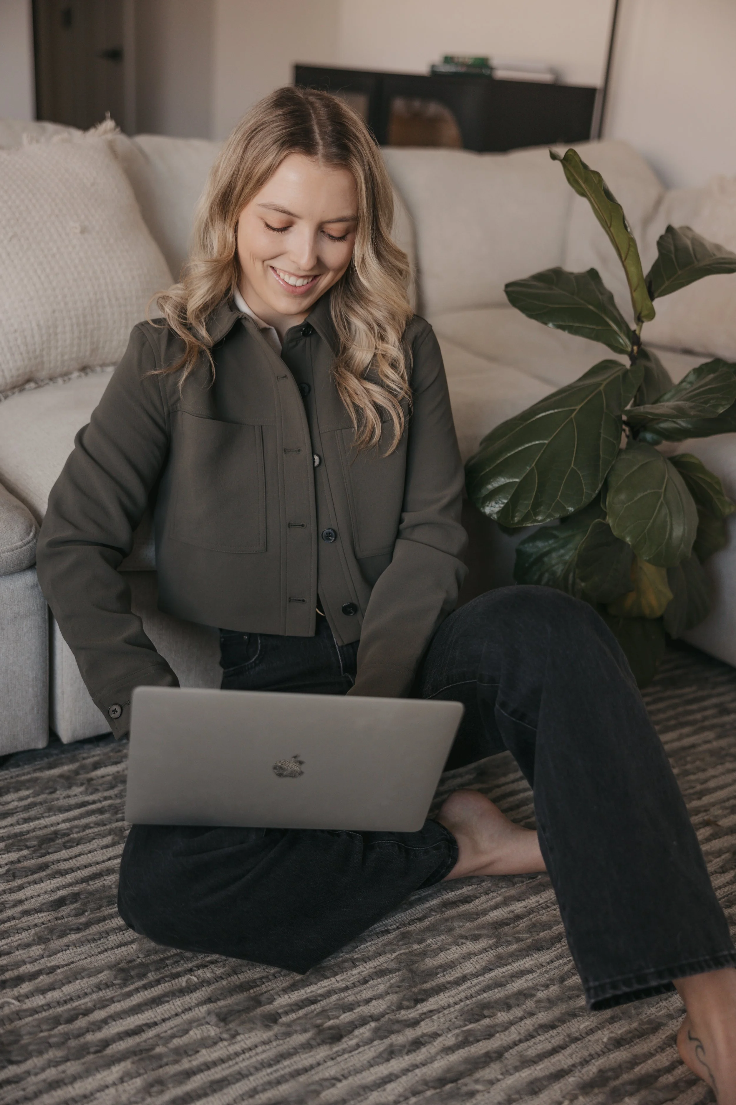 A woman with blonde hair sitting on the floor with a laptop in her lap, smiling and looking down. She is wearing a gray jacket and dark jeans, and she is barefoot. There is a large houseplant next to her and pillows on a couch behind her.