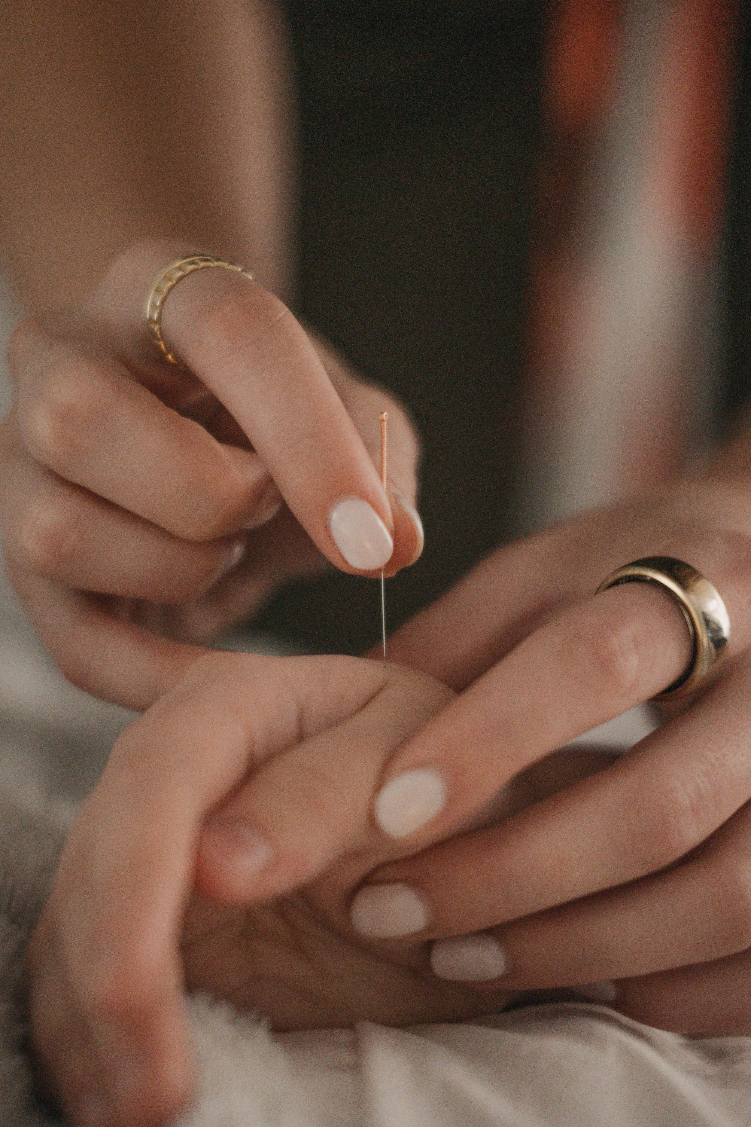 Close-up of hands holding a needle during a sewing or crafting process, with focus on the fingers and needle, rings on fingers, and blurred background.