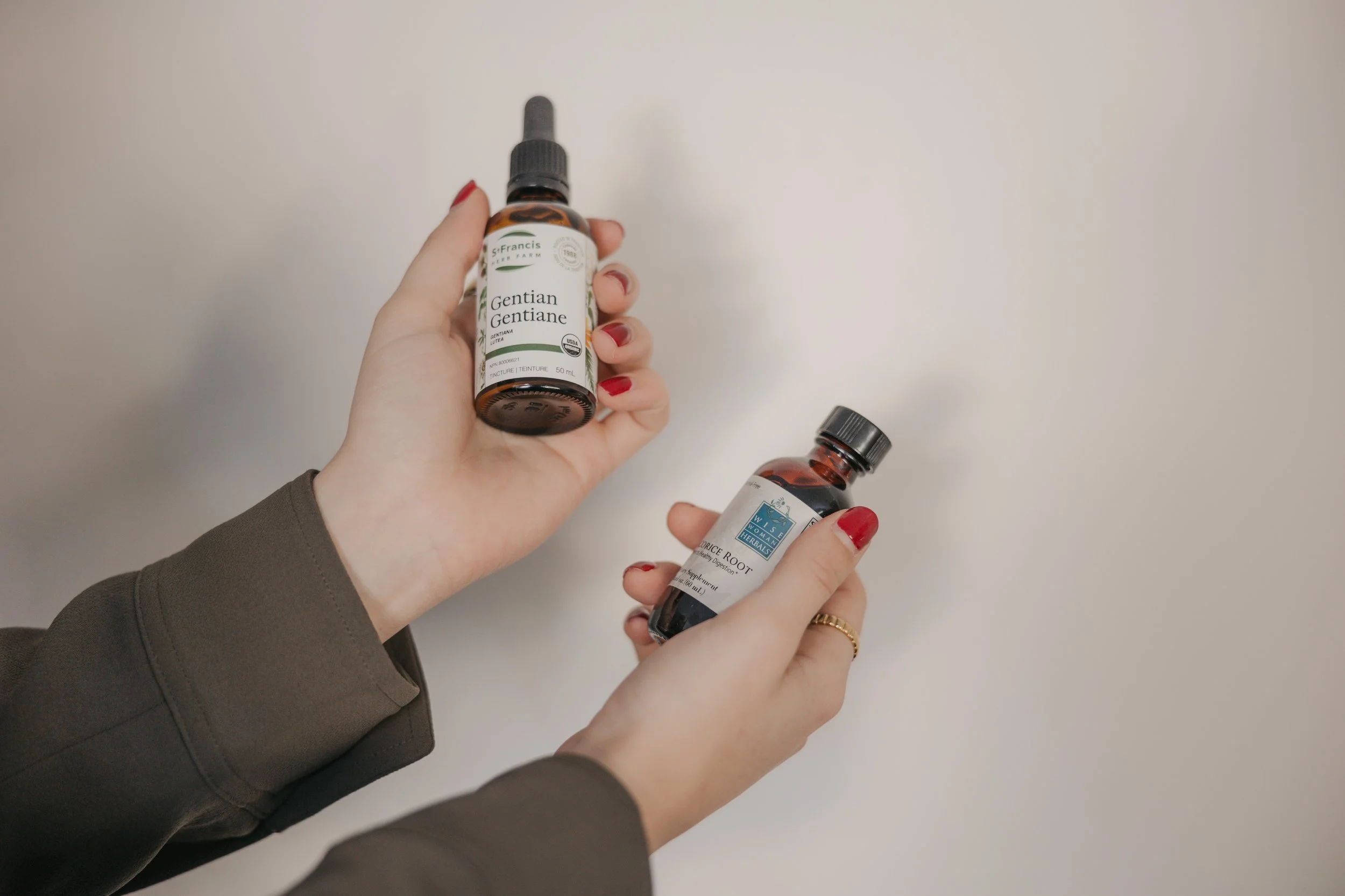 A person with red painted nails and wearing a gold ring on their right hand holds two bottles of herbal liquid supplements against a plain white wall background. One bottle's label reads 'Gentian Gentiane' from St. Francis Herb Farm, and the other is from Wise Woman Herbals.