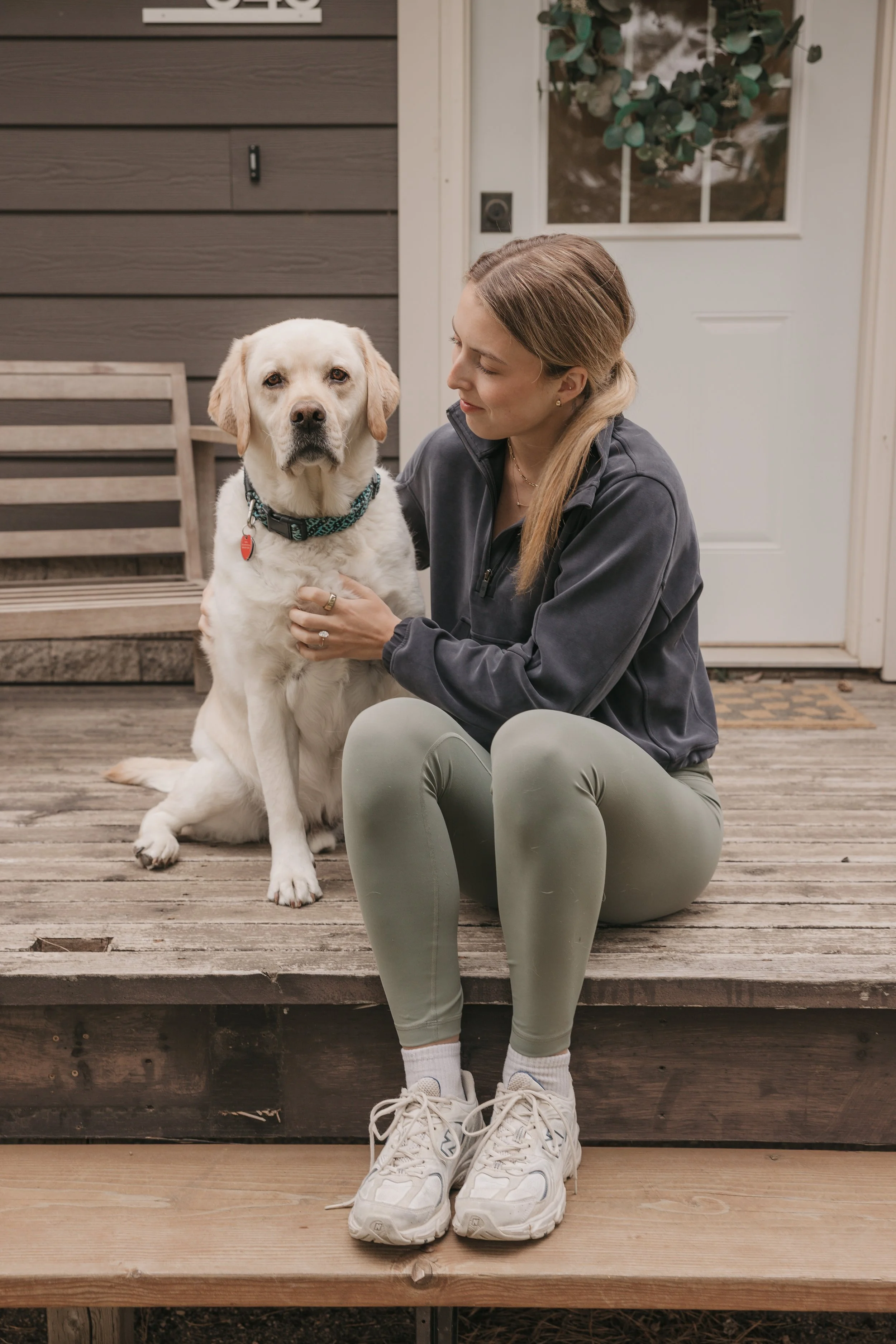 A woman sitting on wooden steps with a Labrador Retriever dog, holding the dog's collar, in front of a house door with a plant visible through the window.