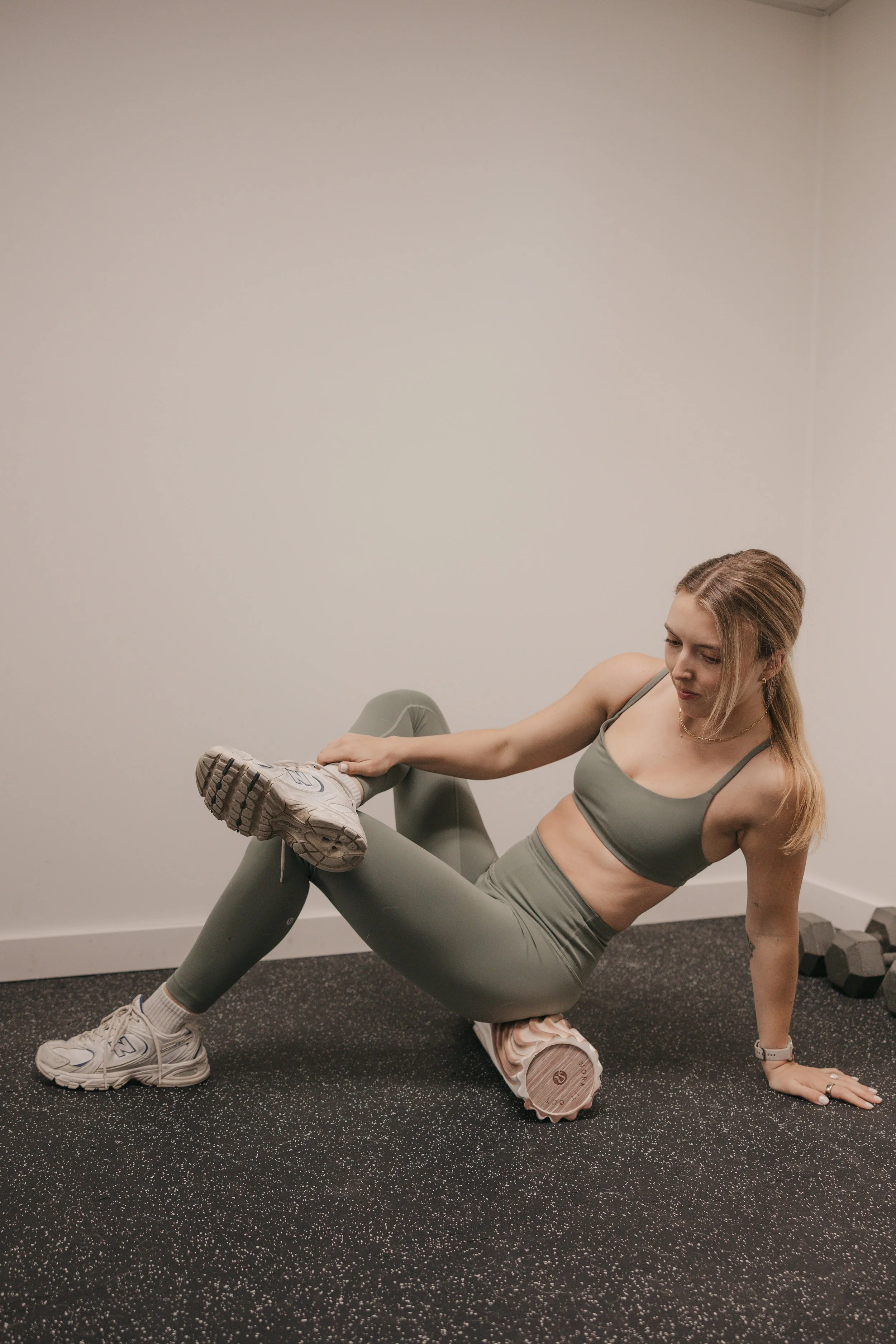 A woman in athletic clothing performs an exercise with her left leg elevated and resting on a foam roller, balancing on her right hand and feet in a gym setting.