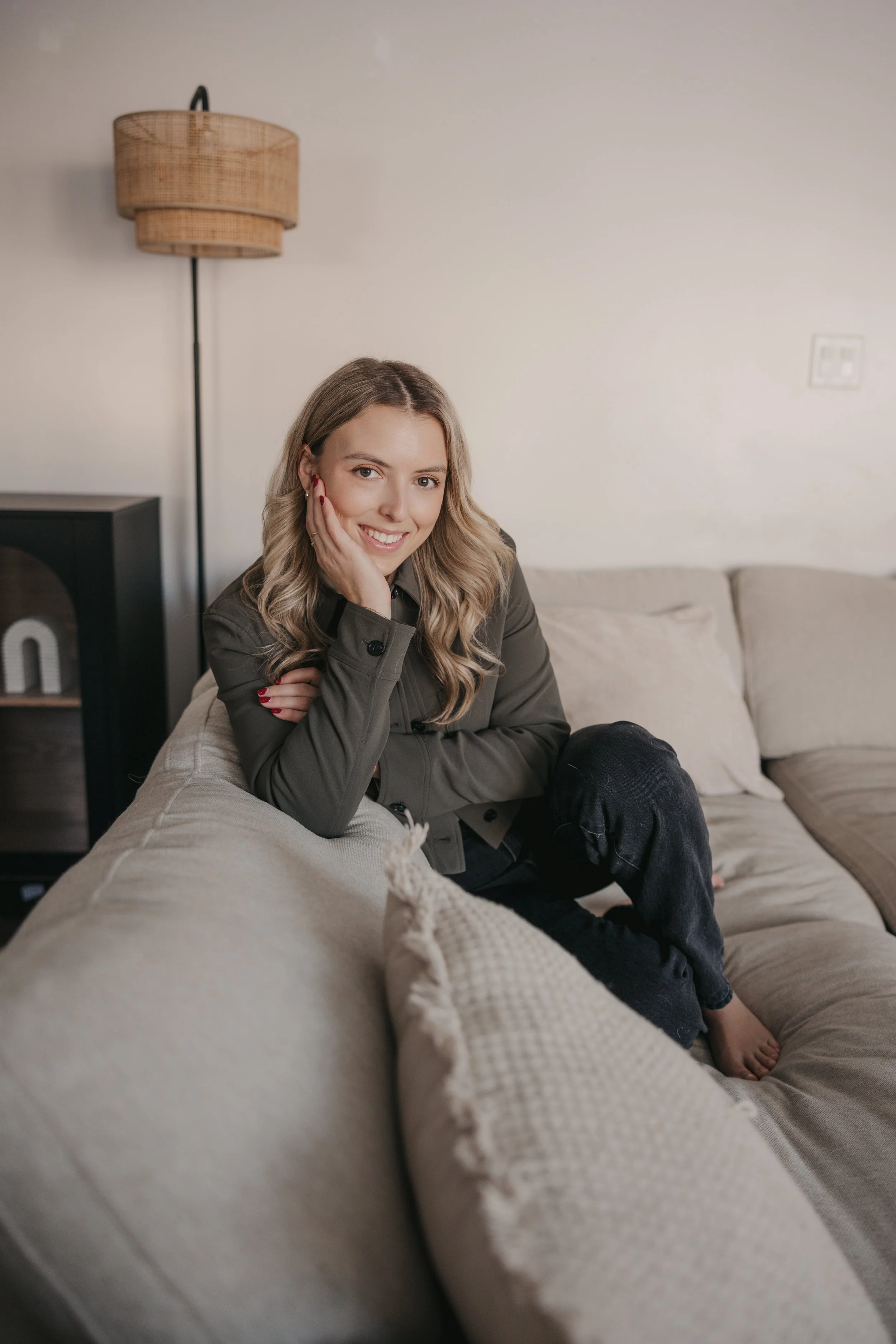 Smiling woman sitting on beige couch in living room, with a cloth on the armrest, in front of a plain wall with a standing lamp and a small side table.