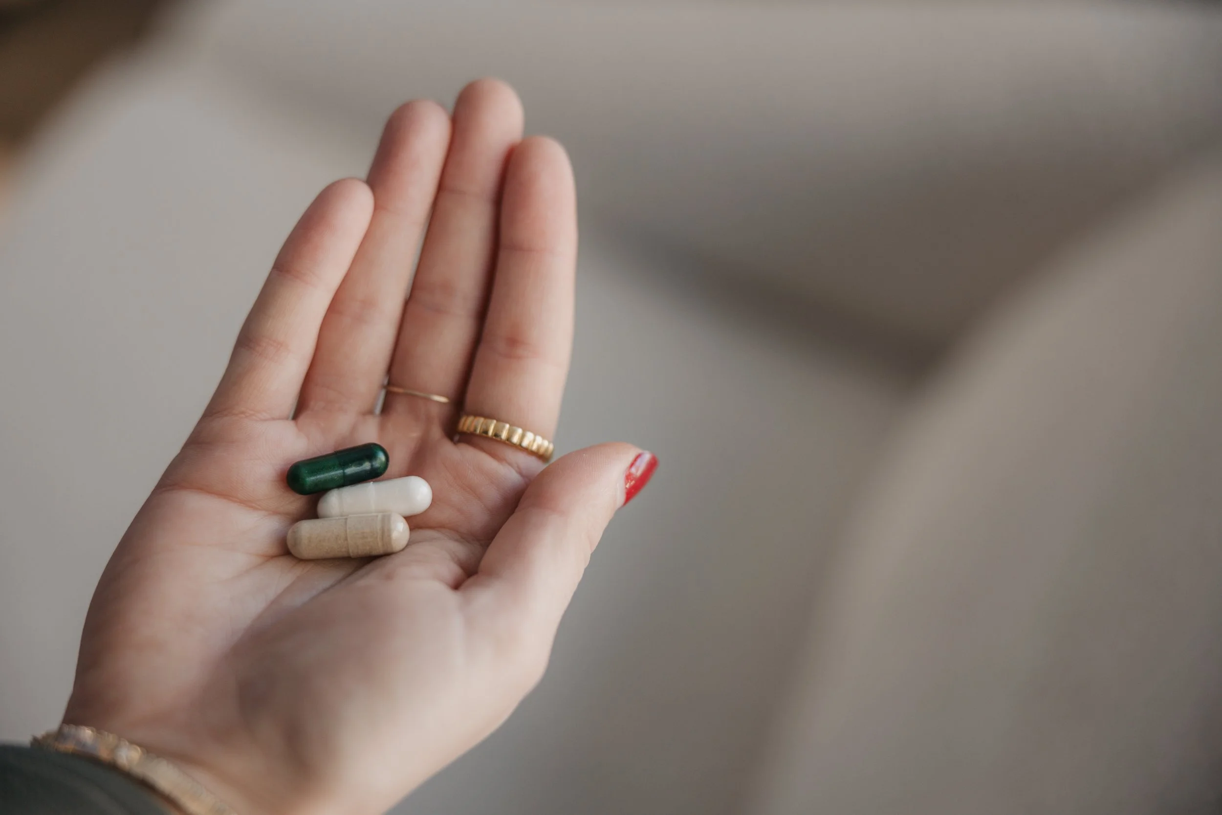 Close-up of a hand holding several different pills and capsules, including green, white, beige, and clear capsules, with a blurred background.