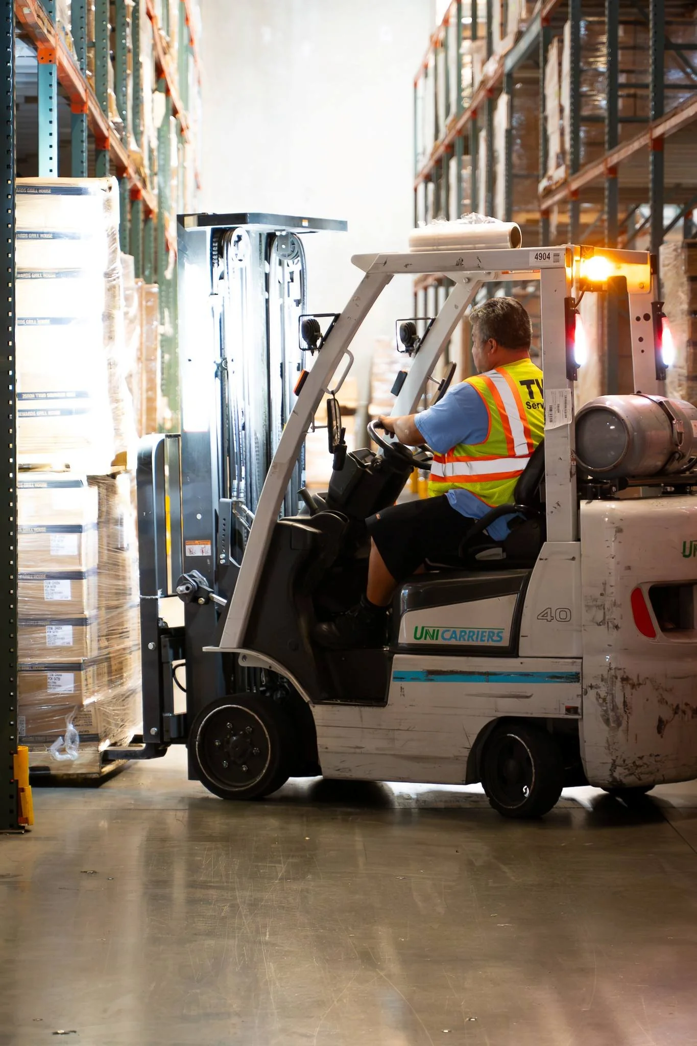 A worker operating a forklift in a warehouse, moving pallets of boxed goods on shelves.