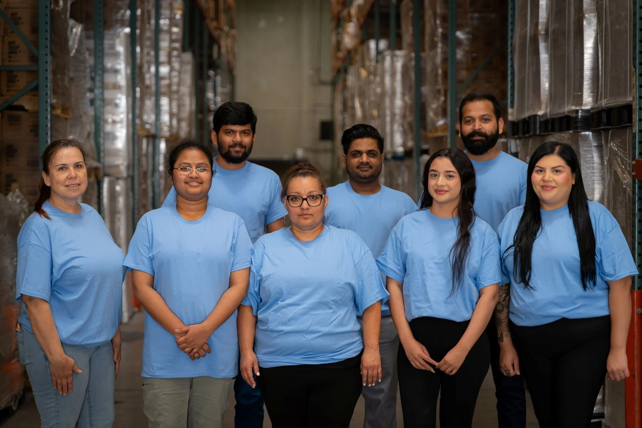Group of eight people wearing blue shirts standing in a warehouse with shelves of boxes behind them.