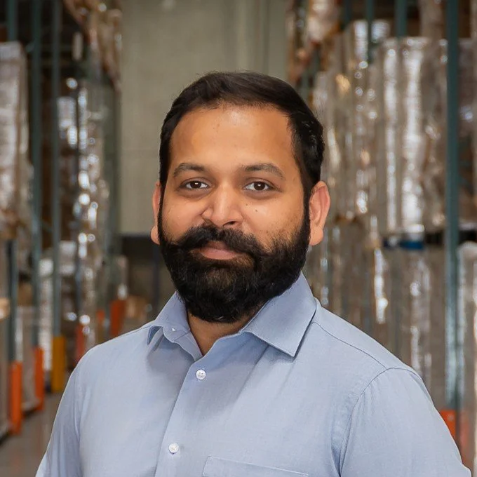 Man with dark hair and beard wearing a light blue button-up shirt standing in a warehouse with shelves and cardboard boxes.