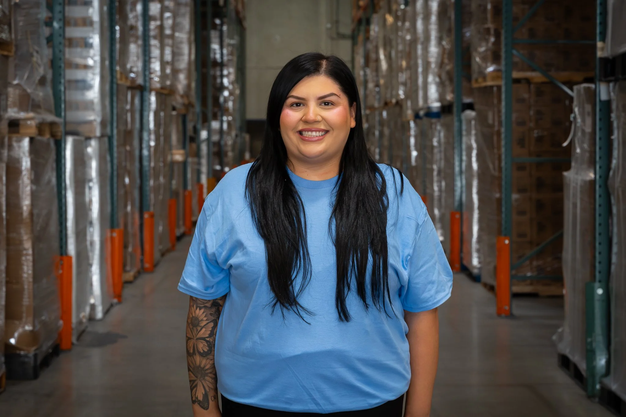 A smiling woman with long black hair and tattoos on her left arm, wearing a light blue t-shirt, standing in a warehouse aisle with metal shelves filled with boxes.