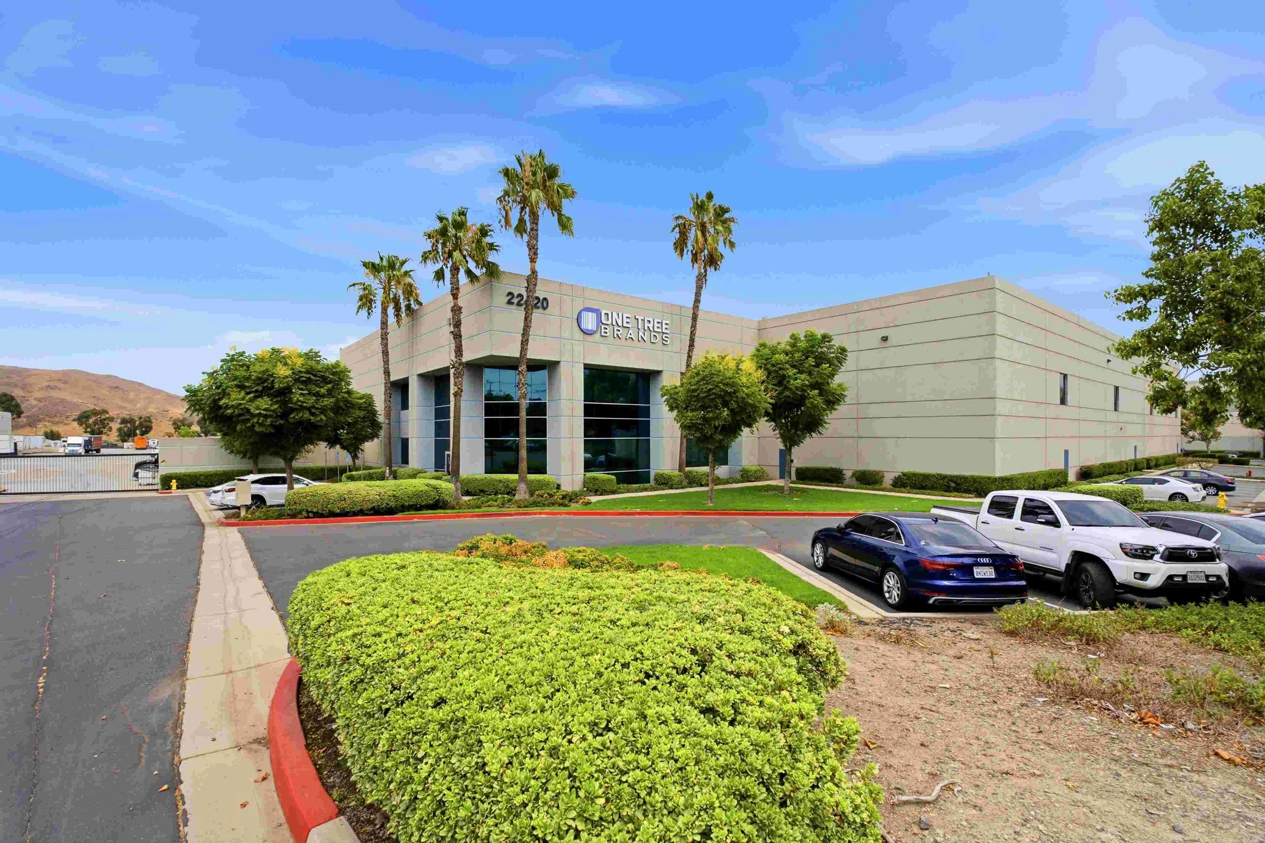 Exterior of a commercial building with a sign that reads 'One Tree Brands', surrounded by trees and parking lot with several cars, under a blue sky with some clouds.