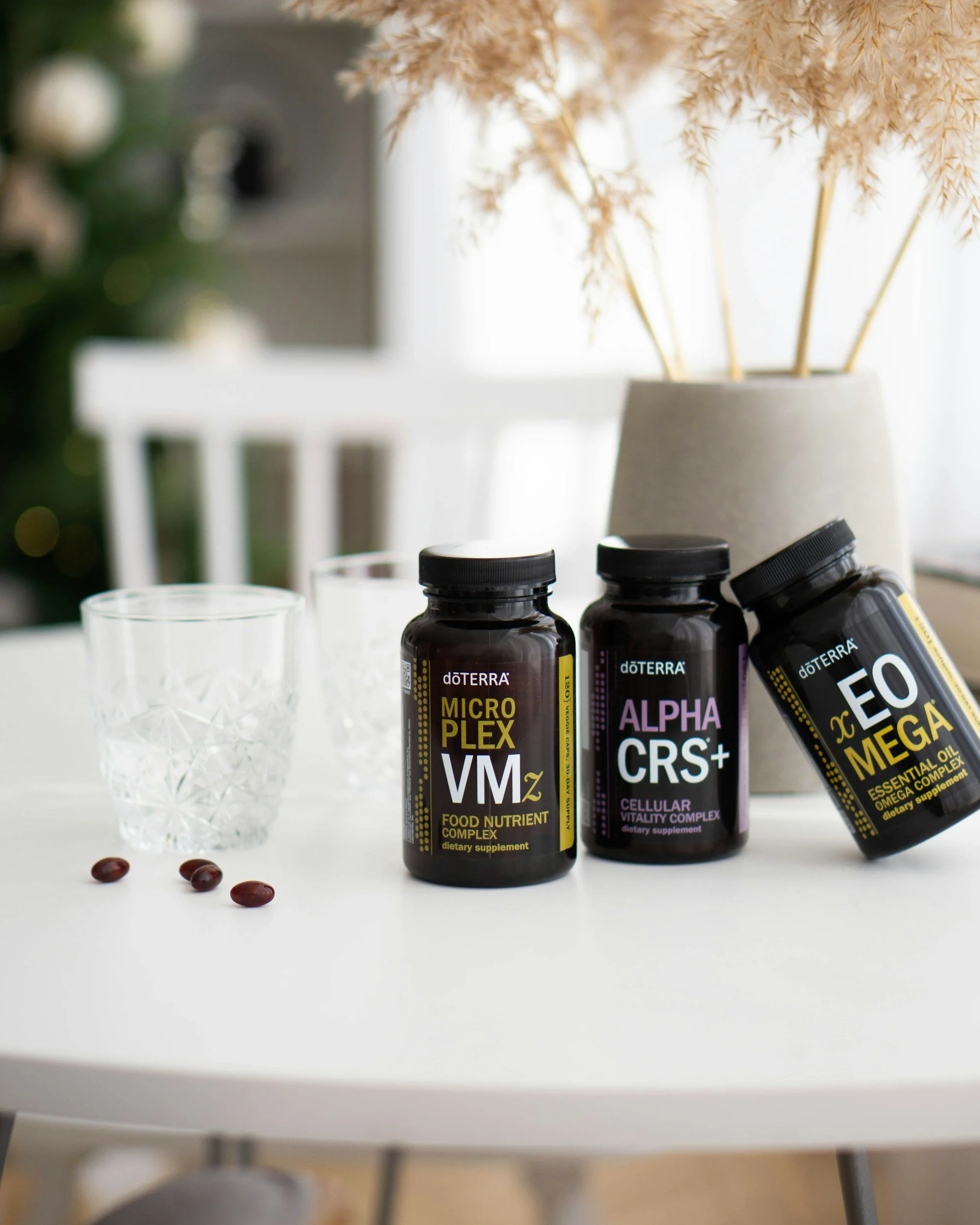 Three bottles of doTERRA essential oils on a white table, with three brown beans and a clear glass of water nearby, and a blurred background of a white chair and a gray vase with beige dried plants.