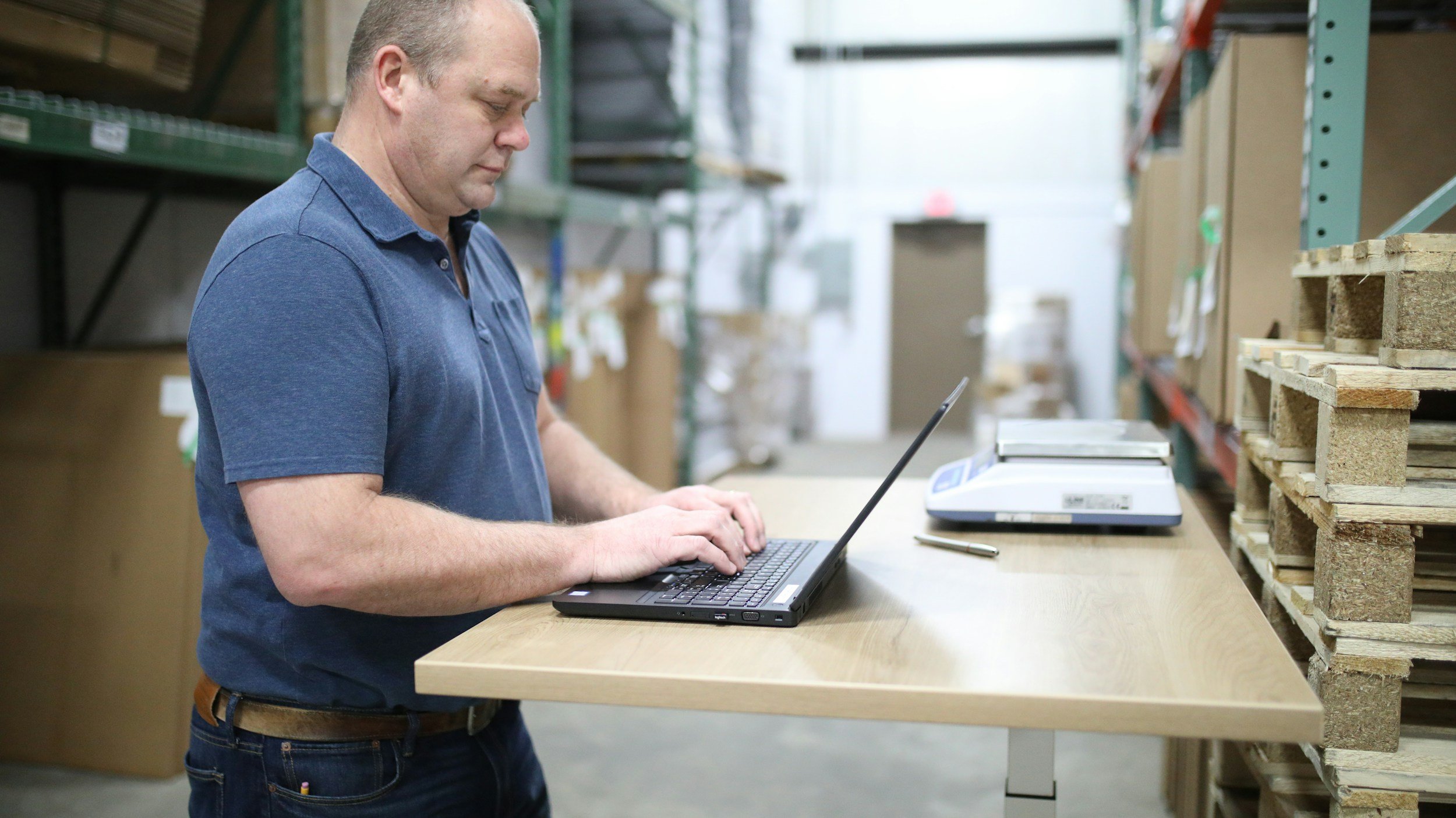 A man in a blue polo shirt working on a laptop at a workbench in a warehouse, with shelves of wooden pallets and boxes in the background.
