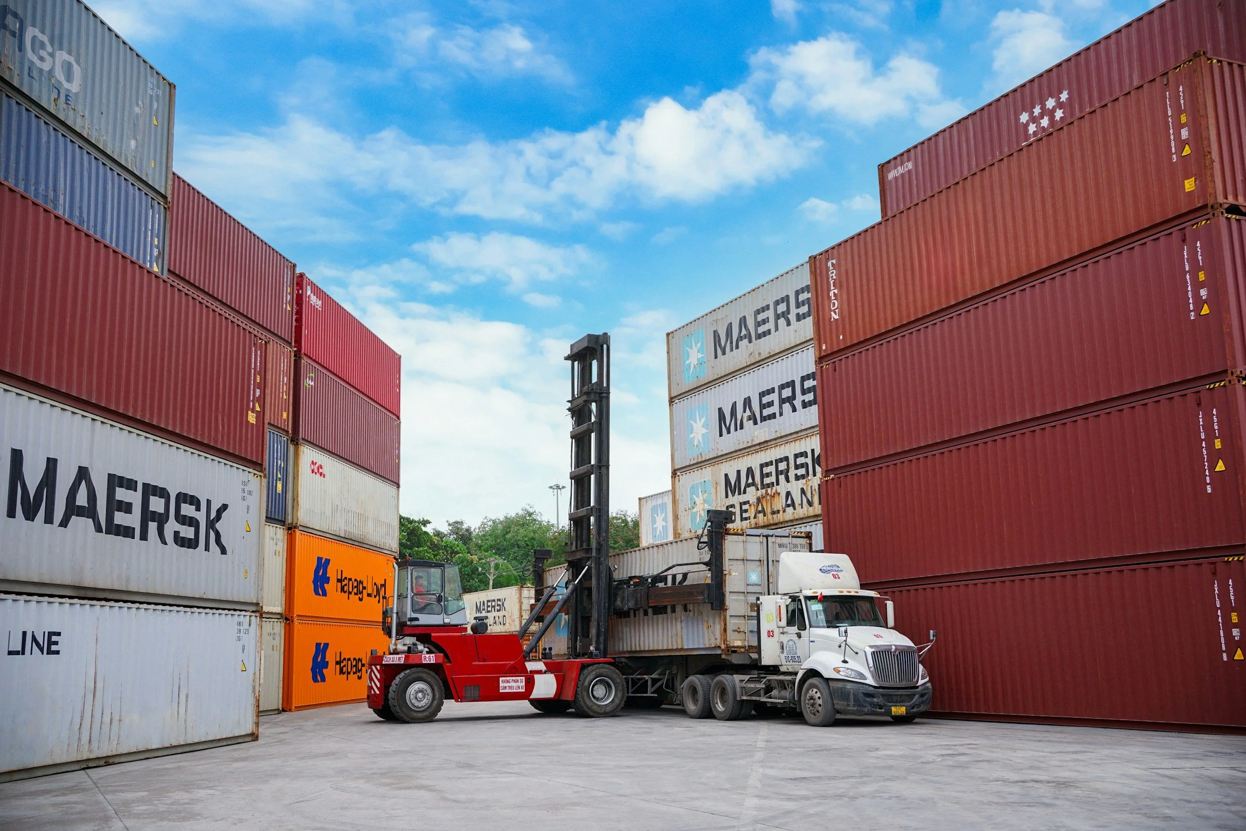 A semi-truck with a forklift loading or unloading containers at a shipping yard. Multiple colorful containers are stacked, with logos like Maersk and Hepa G-Loy.